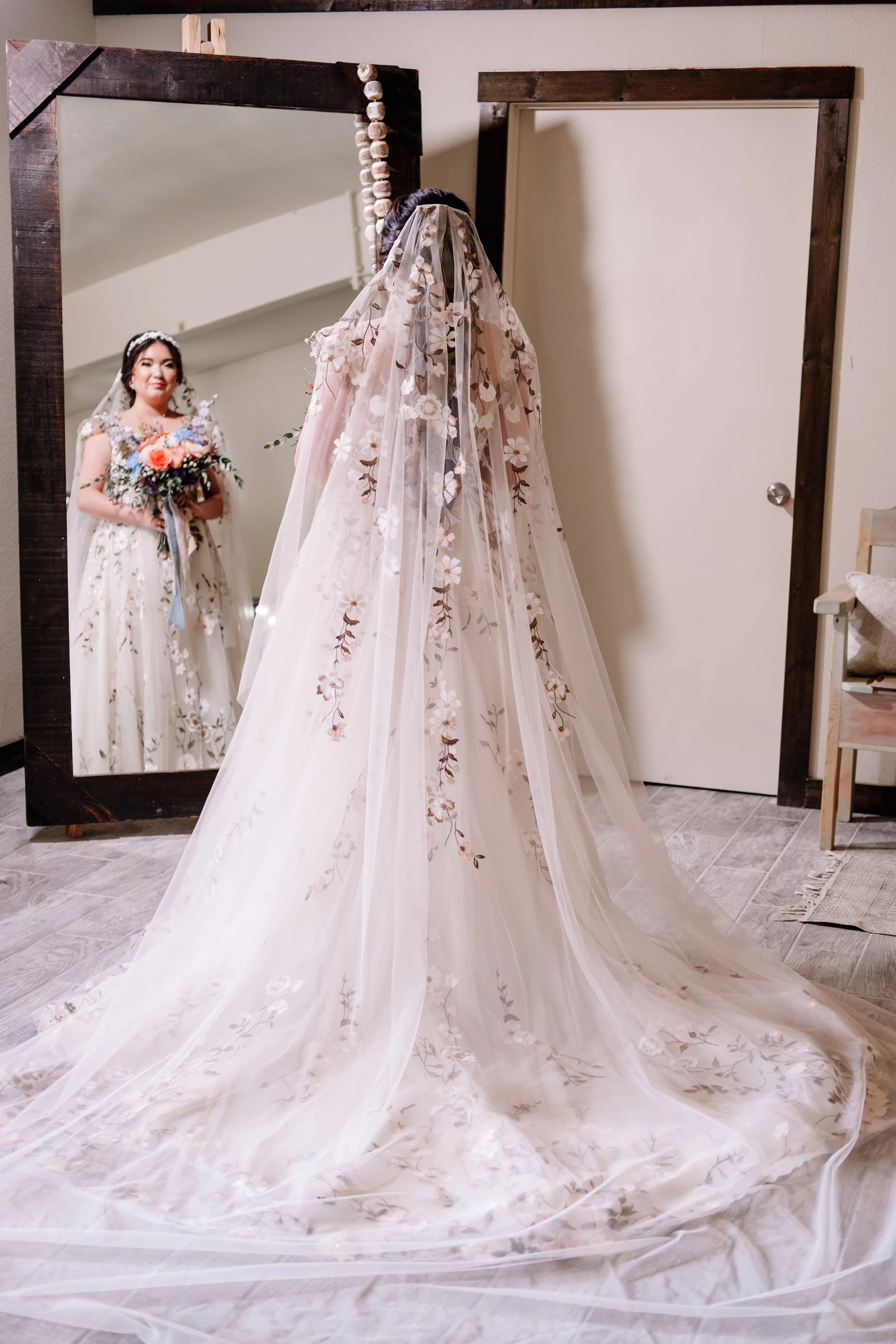 A bride wearing a white wedding dress with floral embroidery, looking at herself in a large mirror, holding a bouquet of flowers.