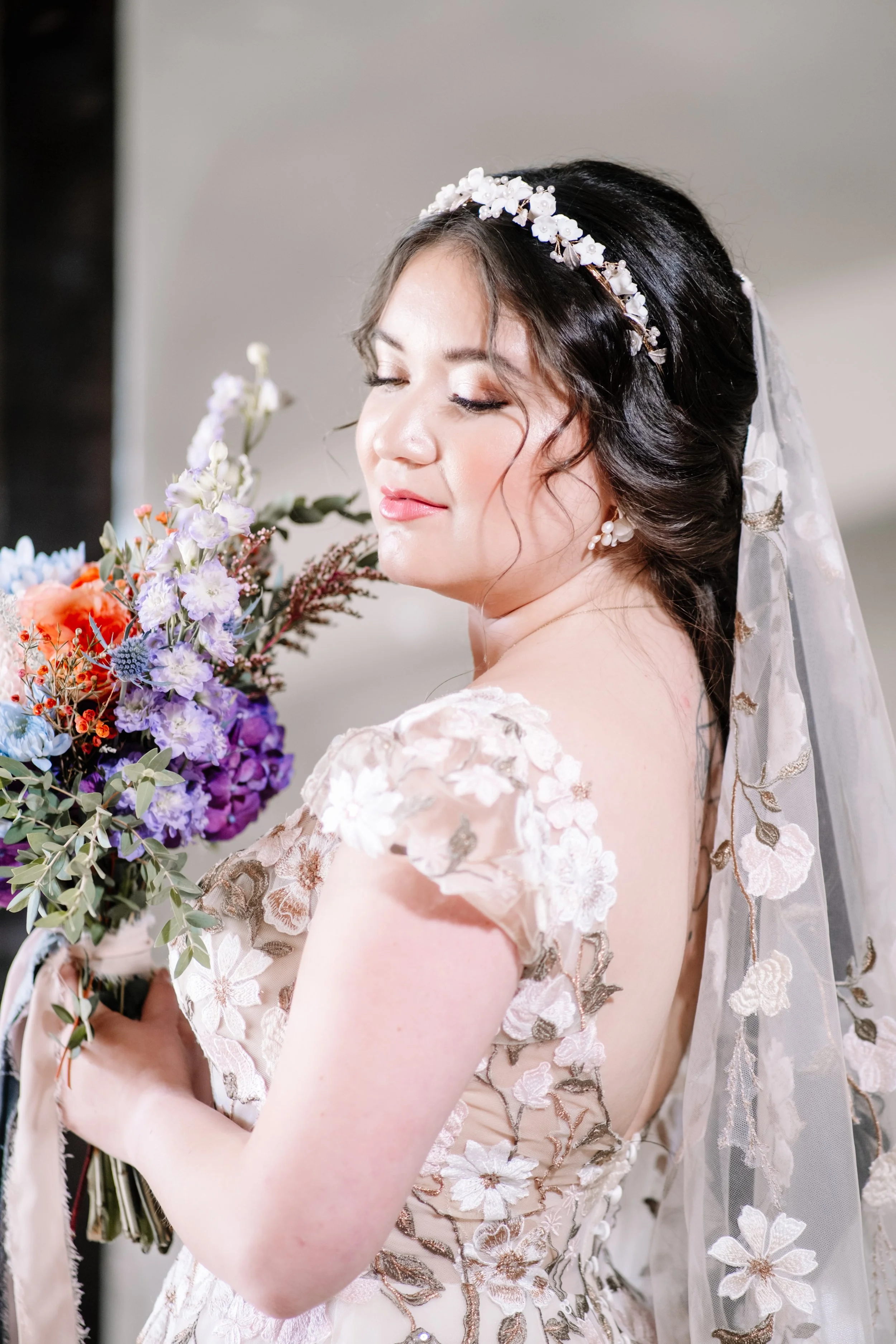 Close-up of a bride in a lace wedding dress with floral embroidery, holding a colorful bouquet, with her eyes closed and a slight smile, wearing a floral headband and veil.