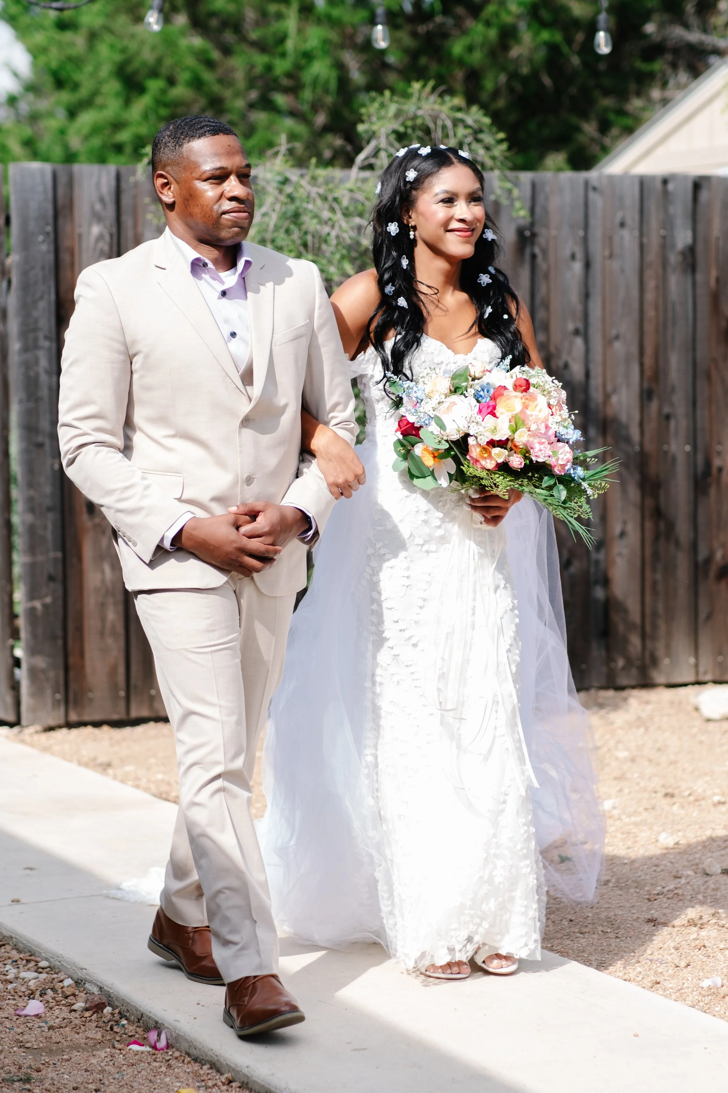 A bride in a white wedding dress holding a colorful bouquet, walking beside a man in a beige suit during an outdoor wedding ceremony.