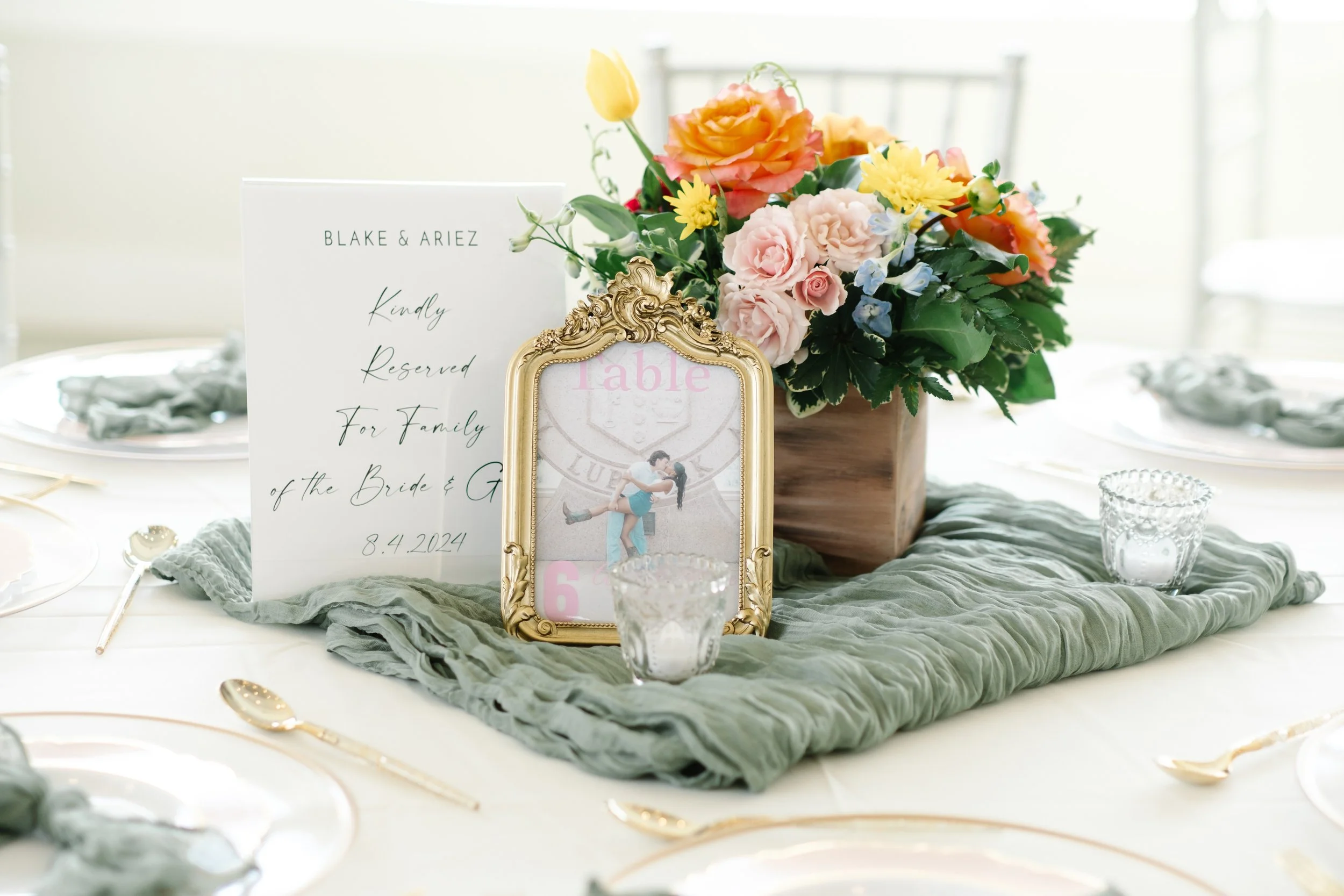 Wedding table centerpiece with a floral arrangement in a wooden box, a gold picture frame with an active photo, and an informational card, all on a table with a green fabric runner and elegant glassware and gold utensils.