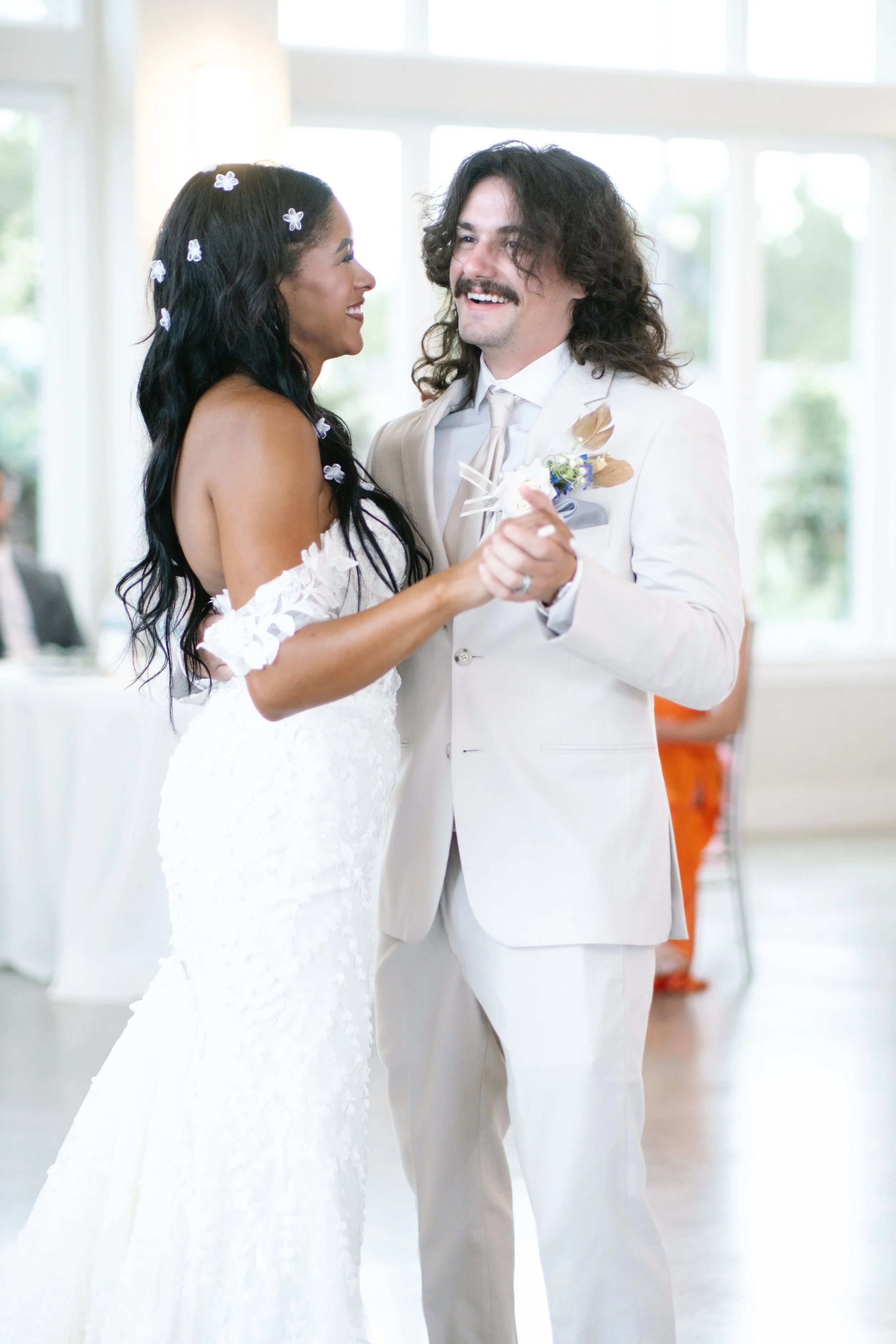 A bride and groom are dancing together at their wedding reception indoors with large windows.