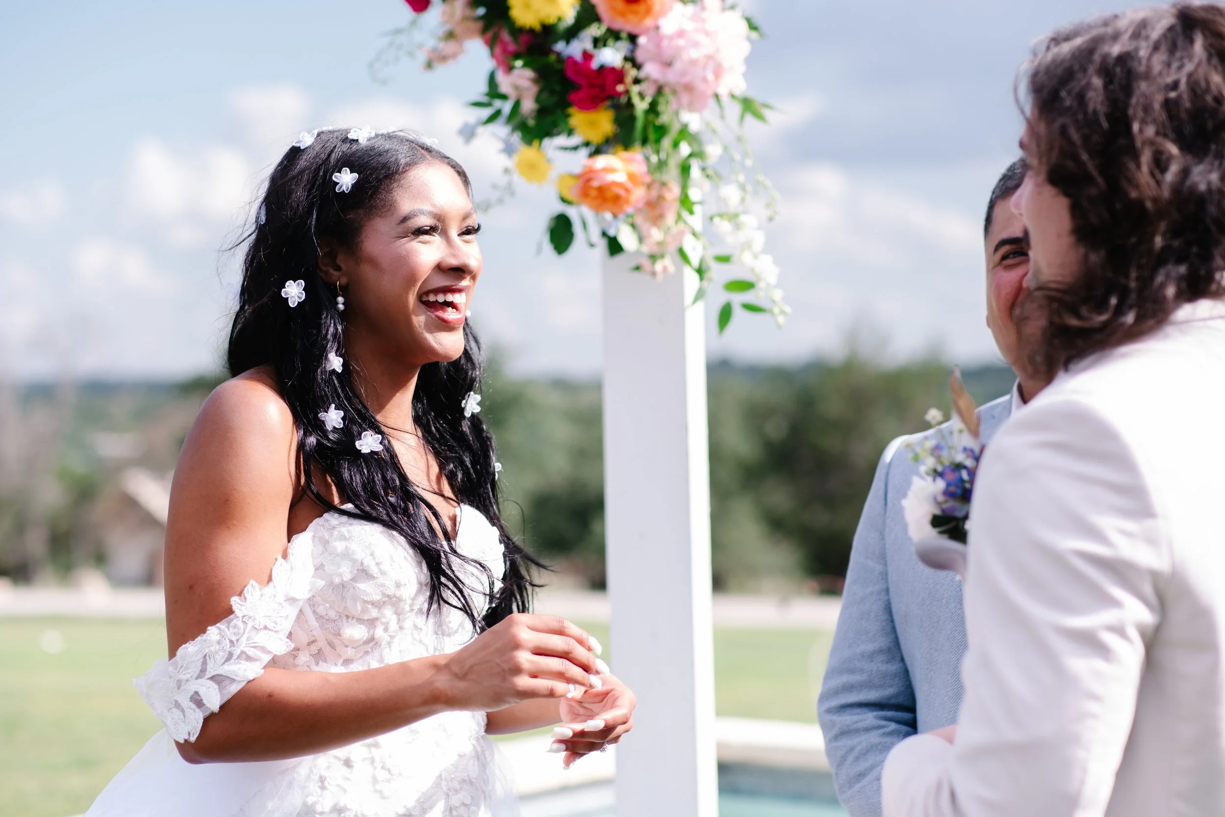 A bride with long dark hair decorated with white flowers, smiling happily during her wedding ceremony outdoors. She is wearing a white lace wedding gown with off-the-shoulder sleeves. A groom and officiant in light-colored suits stand nearby, out of