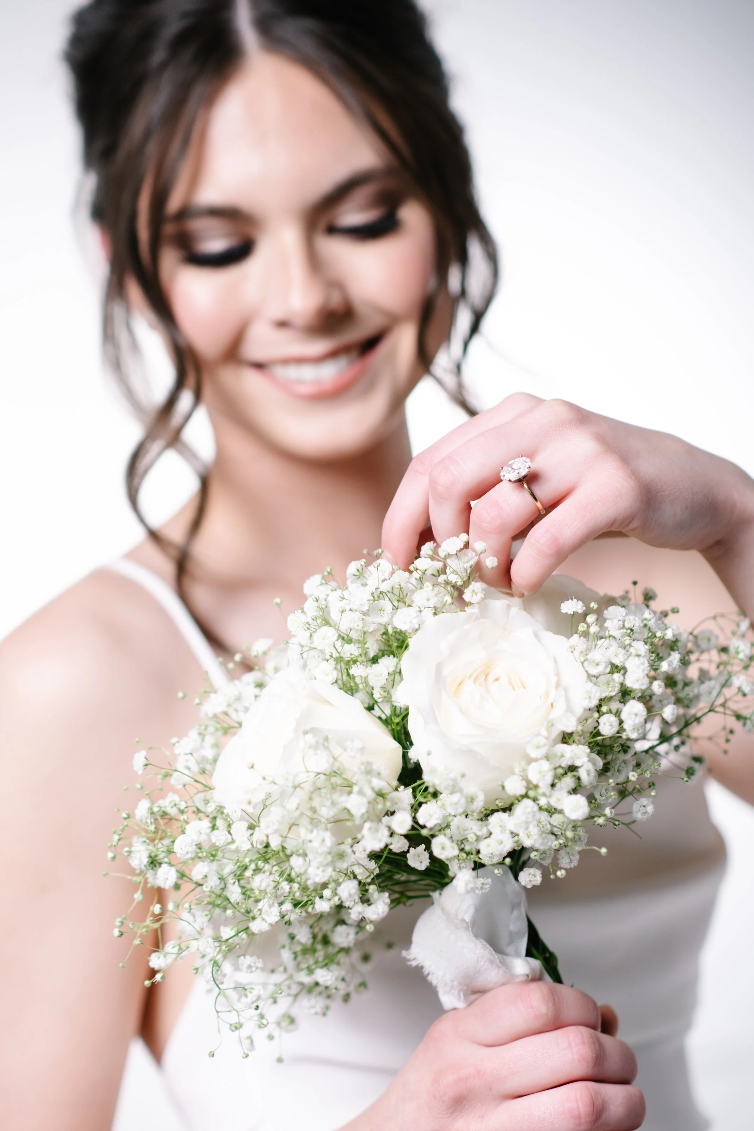 A woman in a white dress holding a bouquet of white roses and baby's breath, smiling with a diamond ring on her finger.