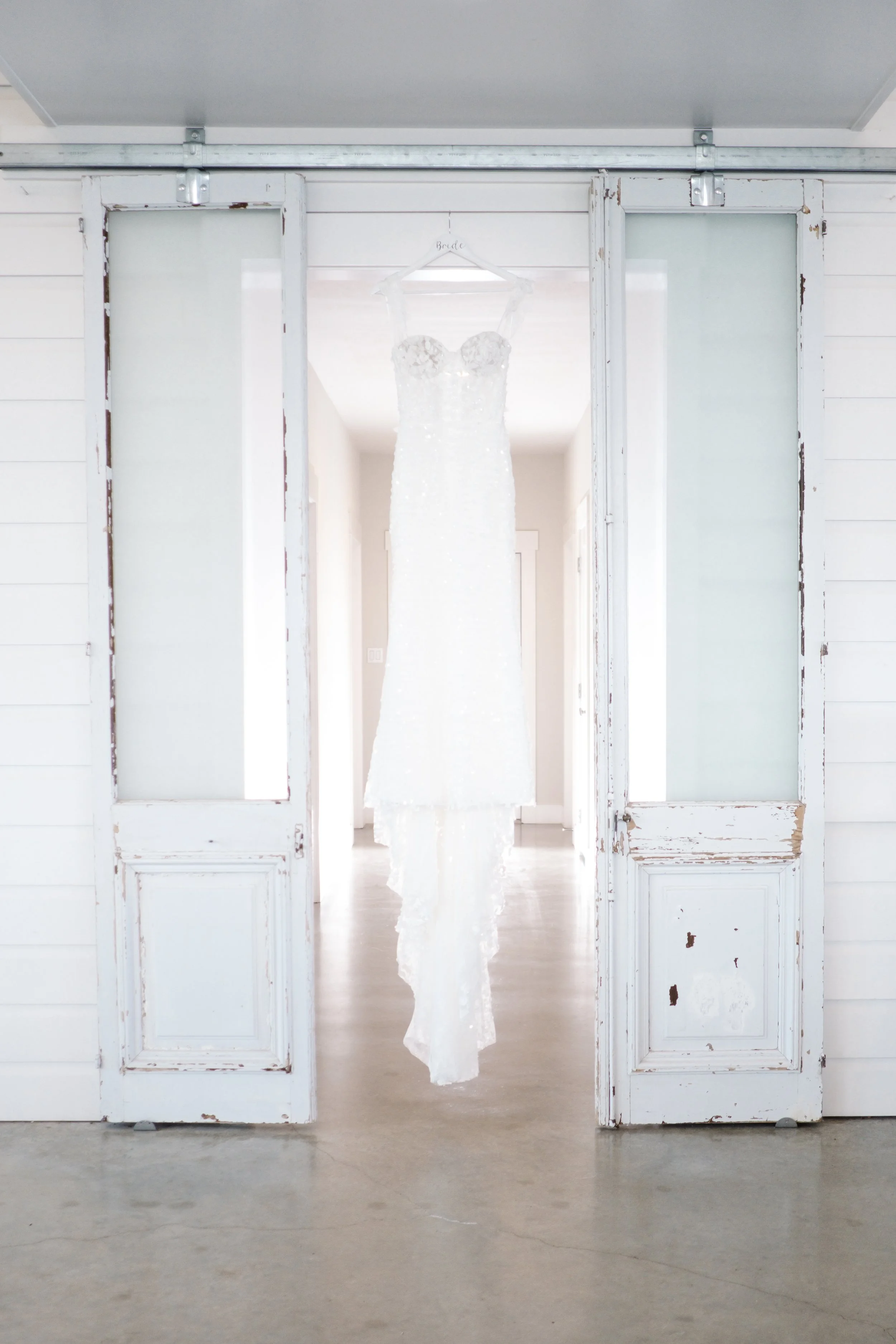 A white wedding dress hanging on a hanger inside a room viewed through rustic white double doors.