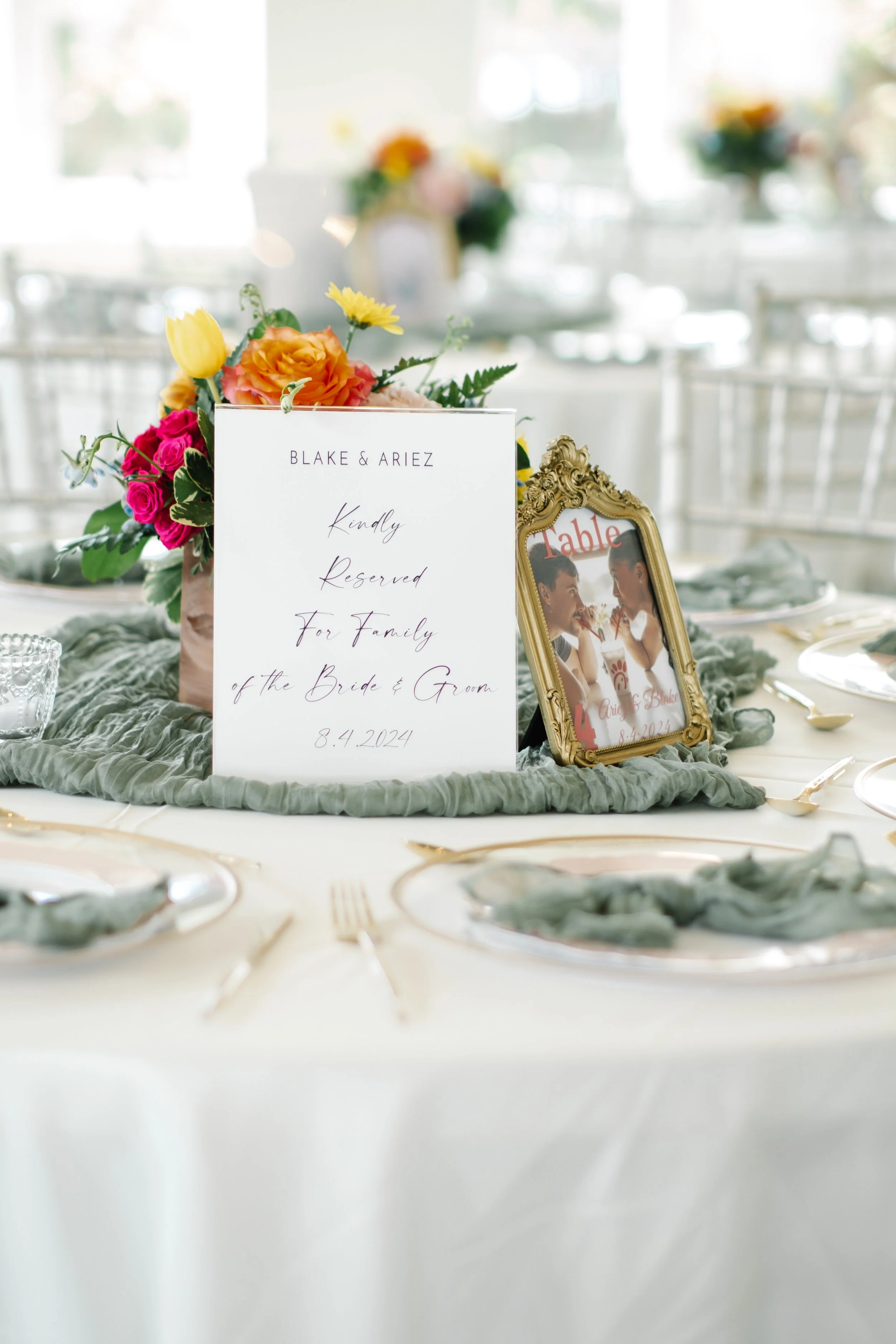 Table setting at a wedding reception with a floral centerpiece, a framed picture of the couple, and a reserved sign for Blake & Ariez dated 8.4.2024.