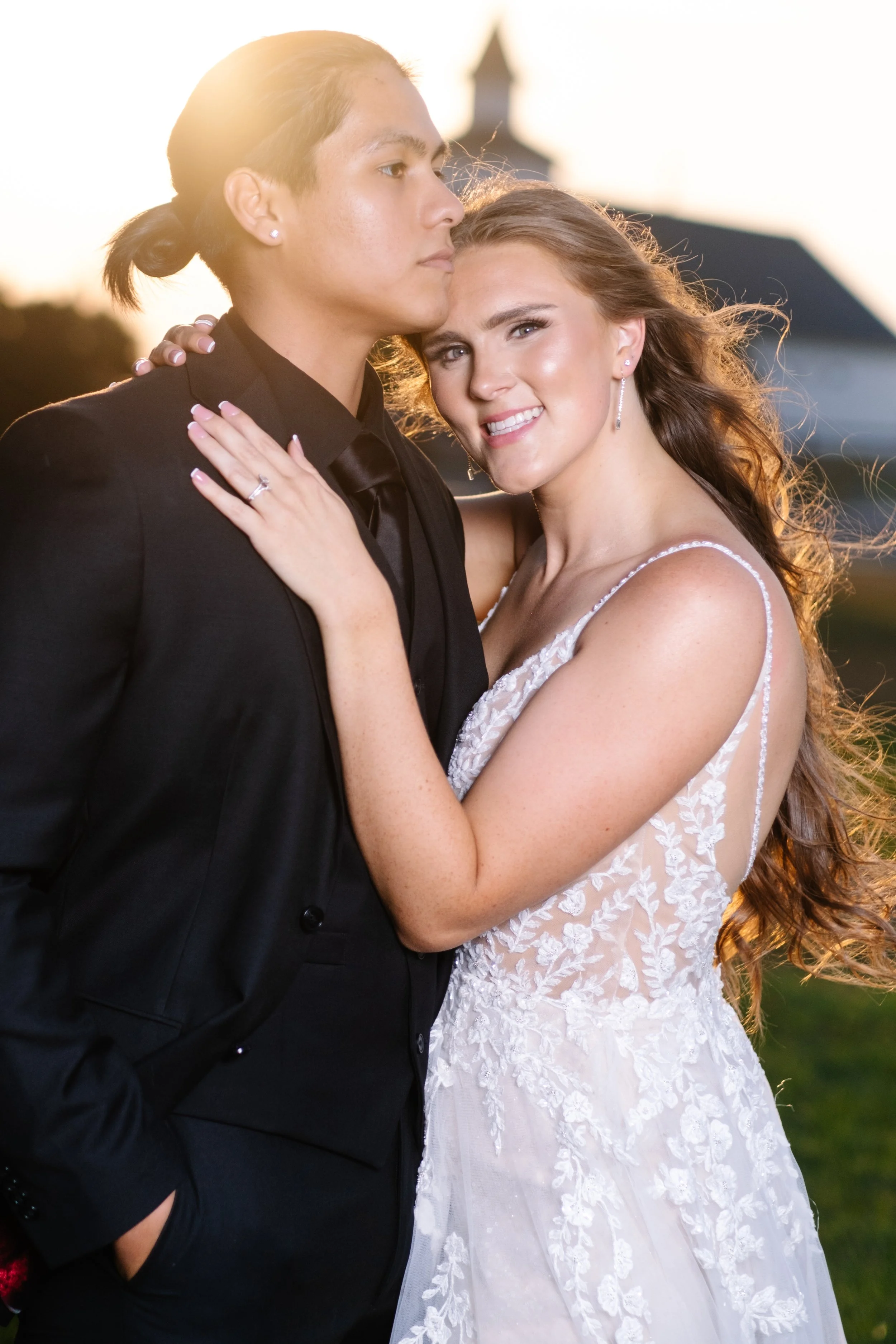 A newlywed couple embracing outdoors during sunset, with the bride smiling and the groom looking serious, both dressed in wedding attire.
