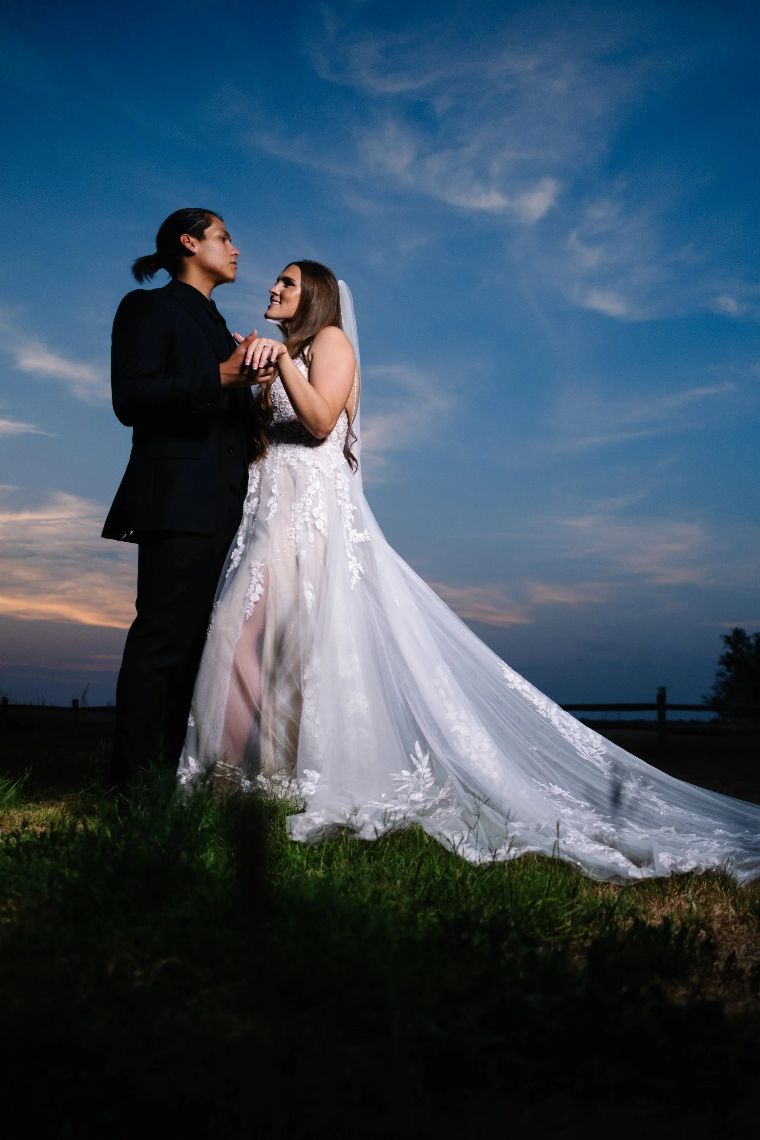 A couple in wedding attire holding hands outdoors during sunset, with the bride wearing a white lace wedding gown and veil, and the groom in a black suit, standing on grass under a sky with streaks of clouds.