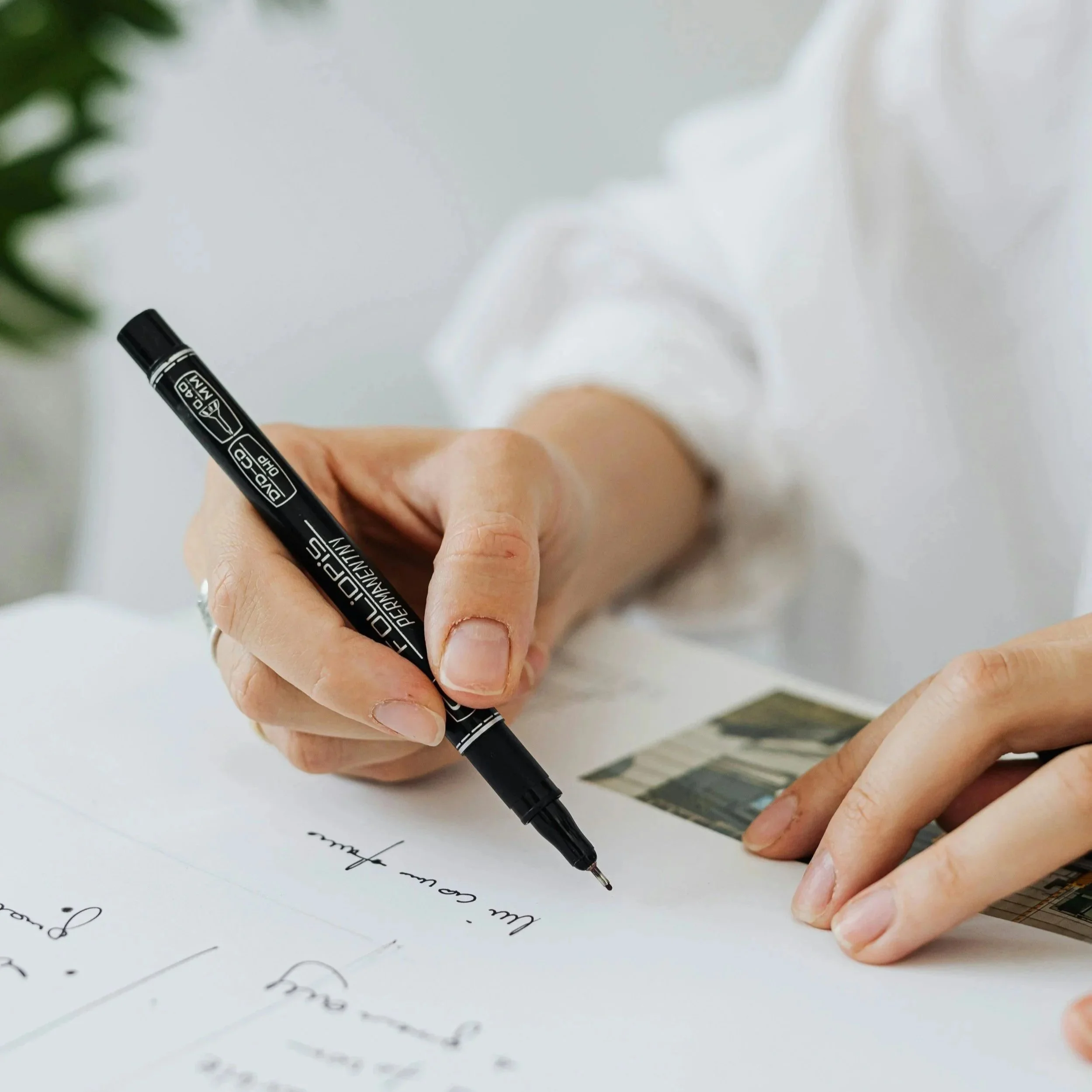 A person writing on a piece of paper with a black marker, holding a photograph and sitting at a white table.