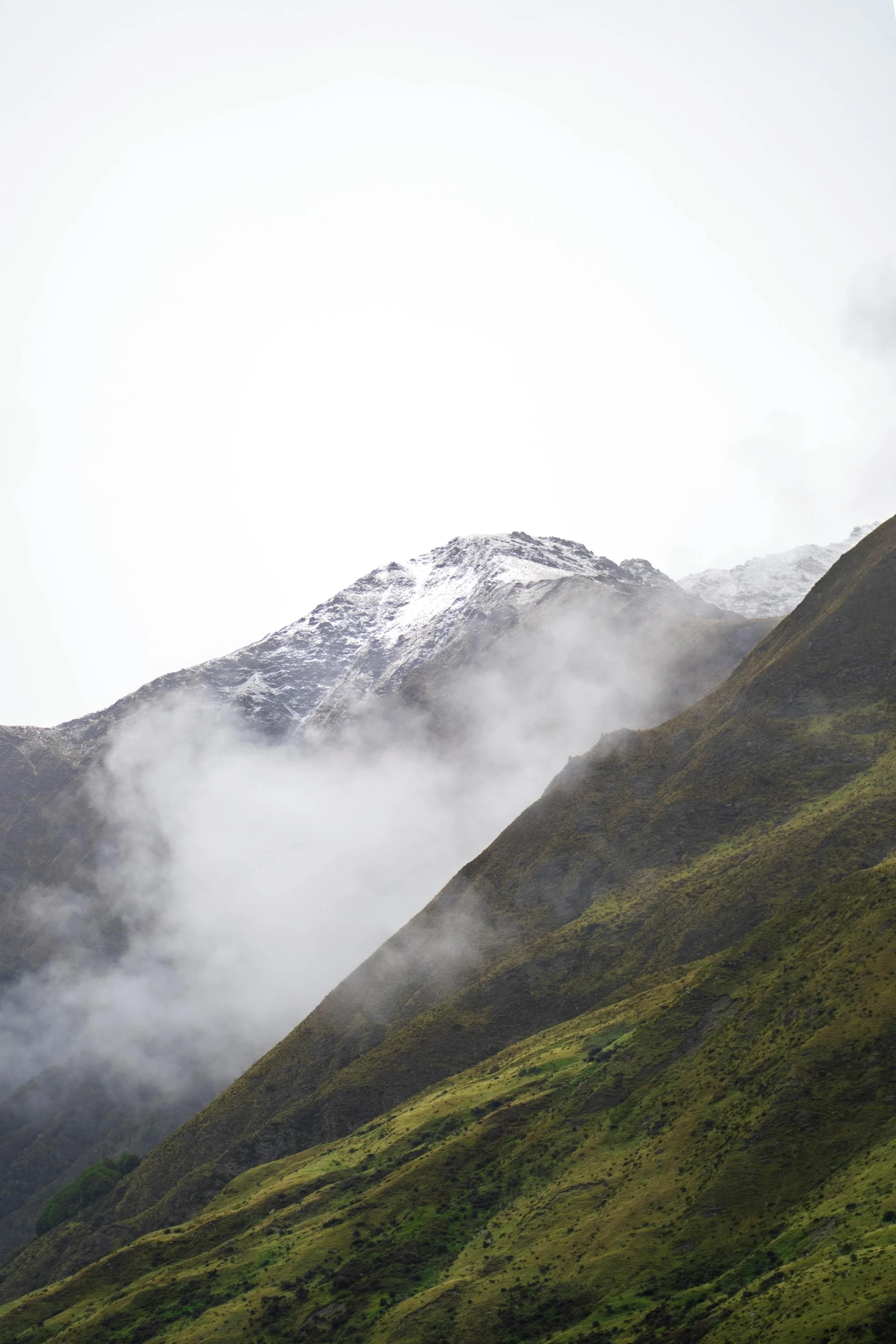 Mountain landscape with green slopes, fog, and snow-capped peaks in the background.