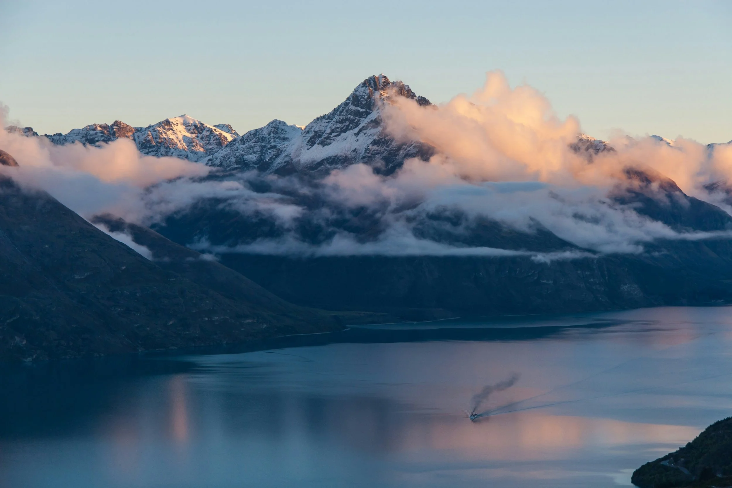 A scenic view of snow-capped mountains with clouds surrounding their peaks, a calm body of water in the foreground, and a boat leaving a trail on the water.