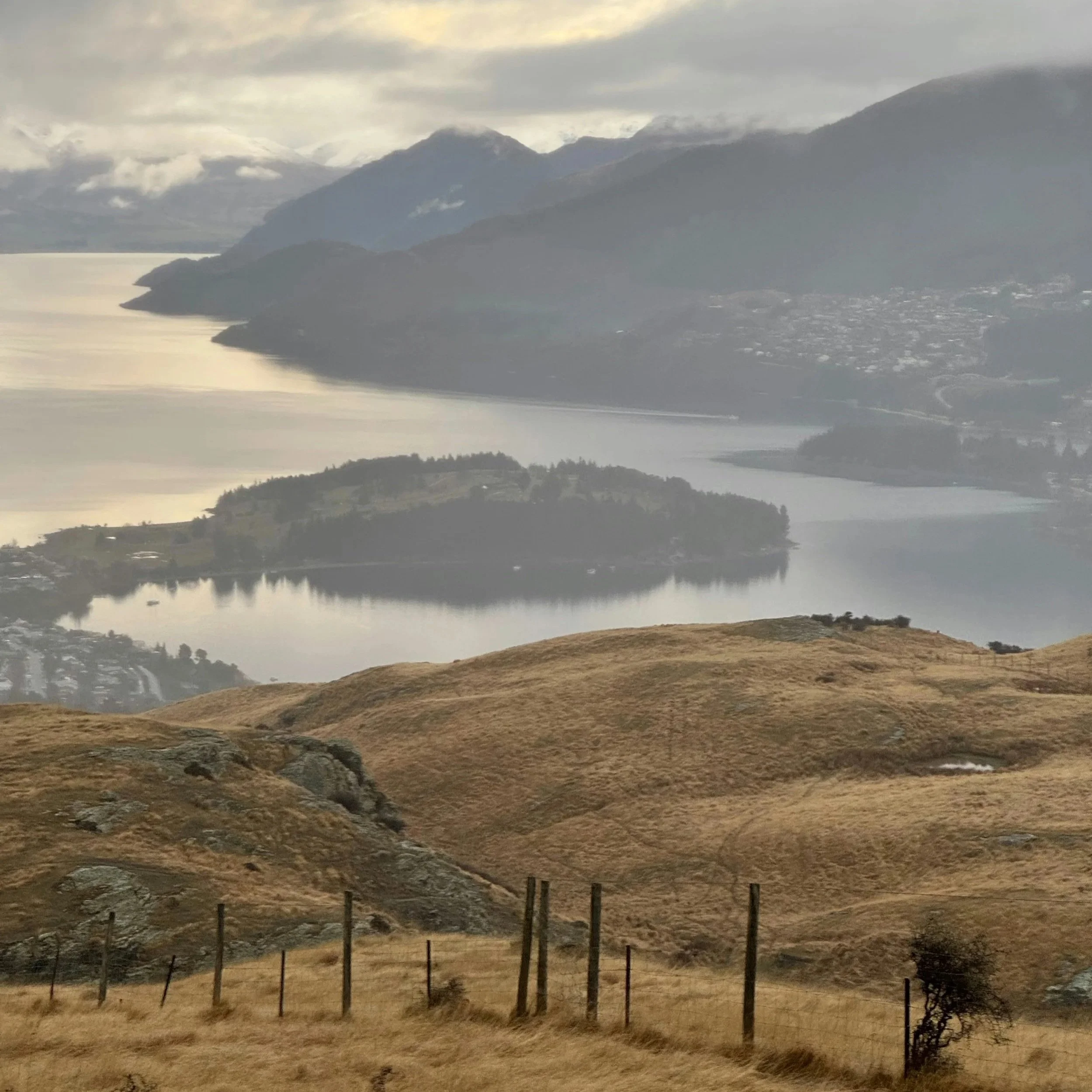 A scenic landscape view of rolling brown hills leading down to a calm body of water with islands and mountains in the background, under a cloudy sky.