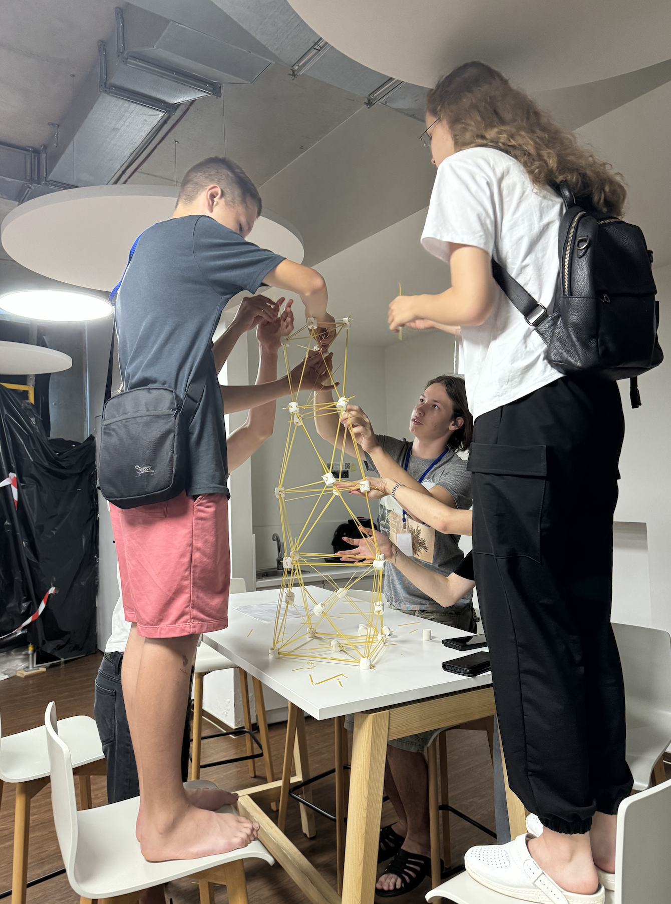 Three young people and a woman assembling a large geometric model made of yellow sticks and white joints on a white table in a modern room.