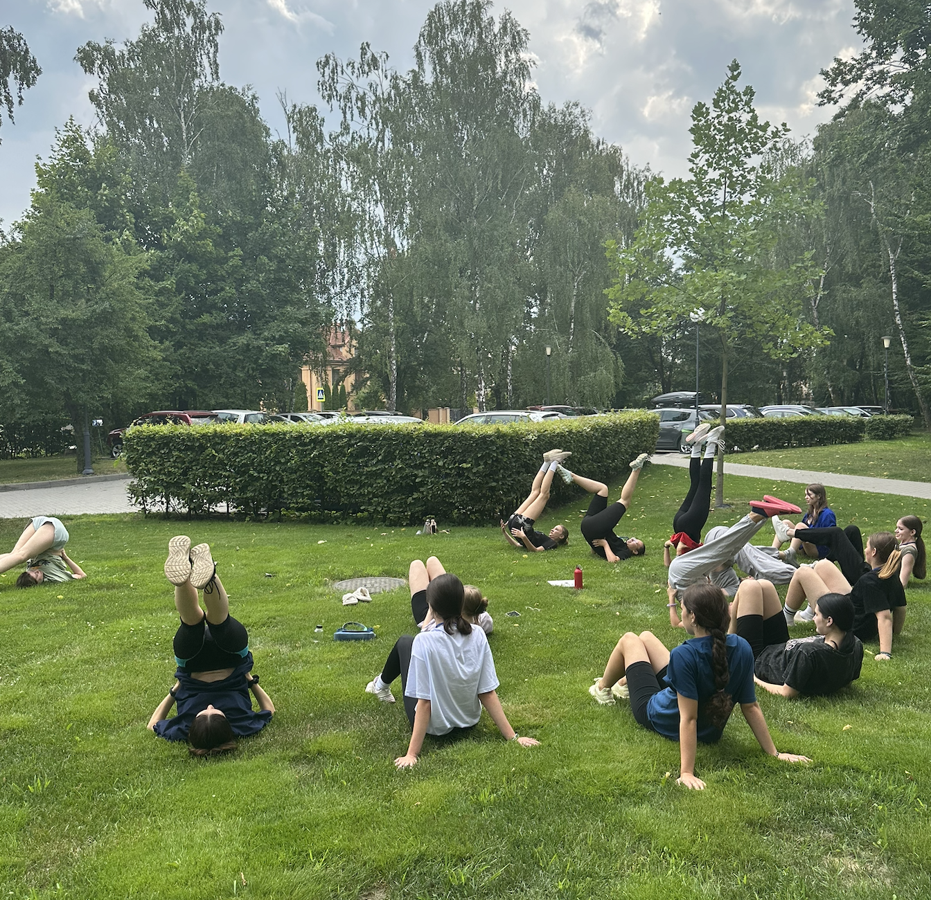 Group of young people exercising outdoors on a grassy area, some doing headstands and others sitting or lying down, with trees and parked cars in the background.