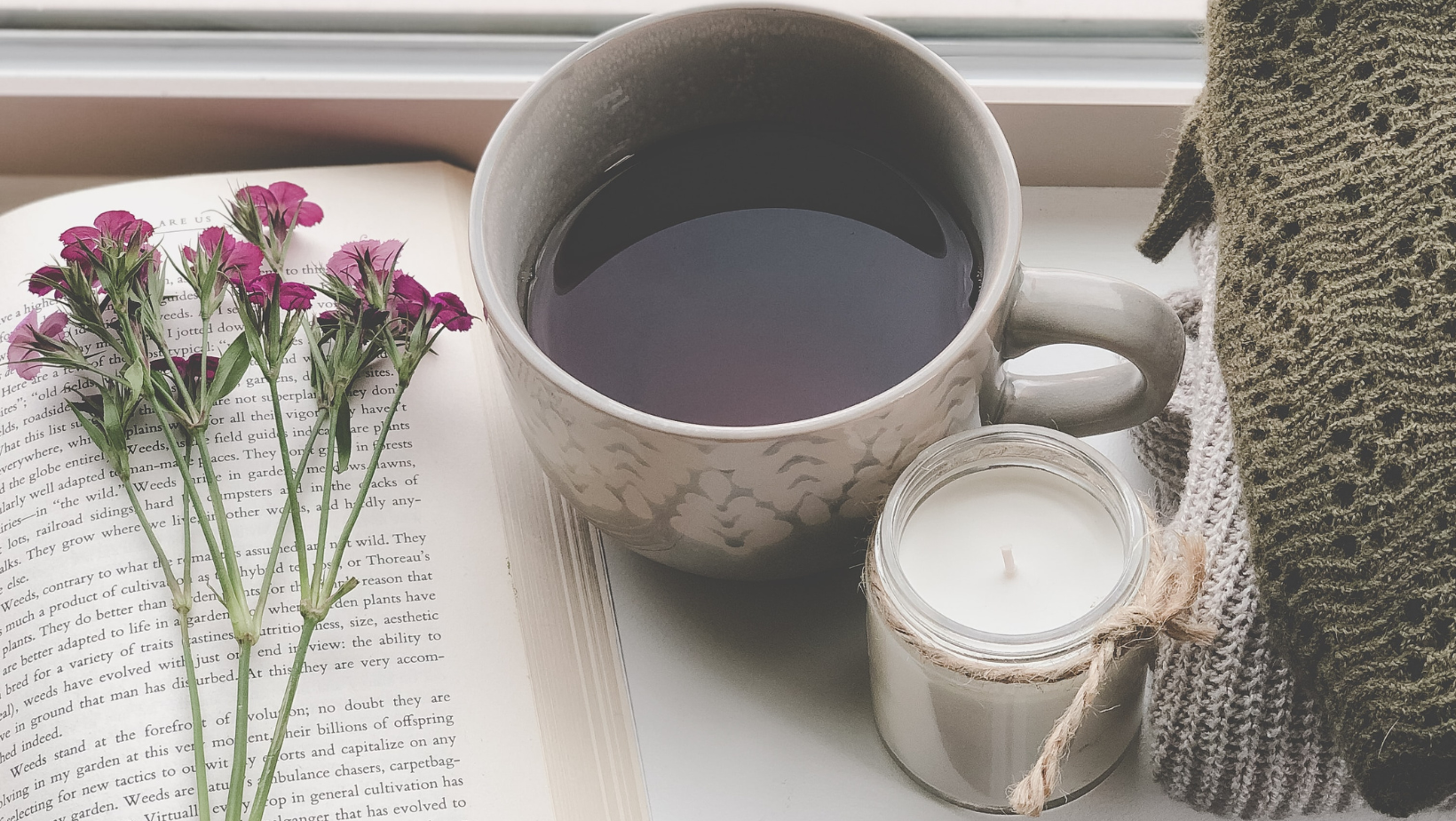 A cozy scene with a book, a bunch of purple flowers, a mug of coffee, a lit white candle in a glass jar, and a gray knitted blanket on a windowsill.