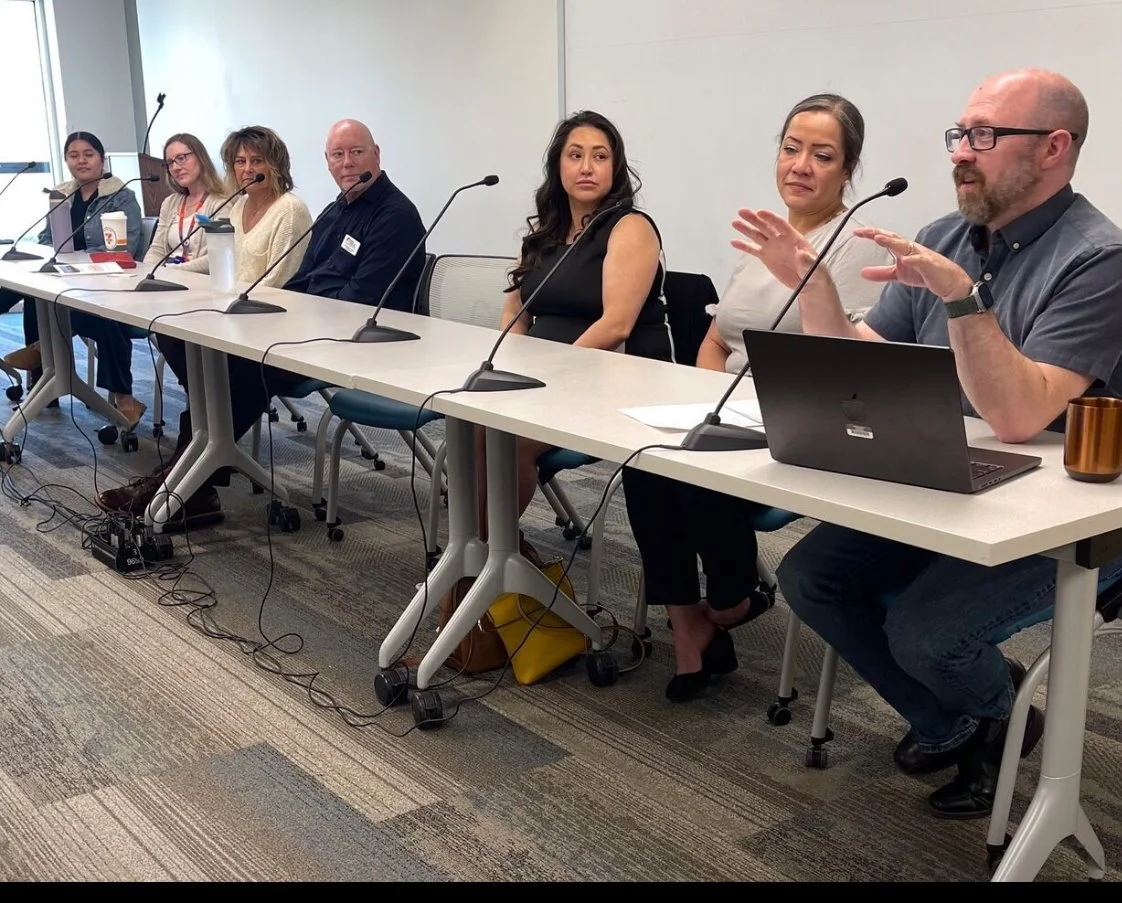 A panel discussion with seven diverse individuals sitting at a long table with microphones in front of each person, in a modern conference room.