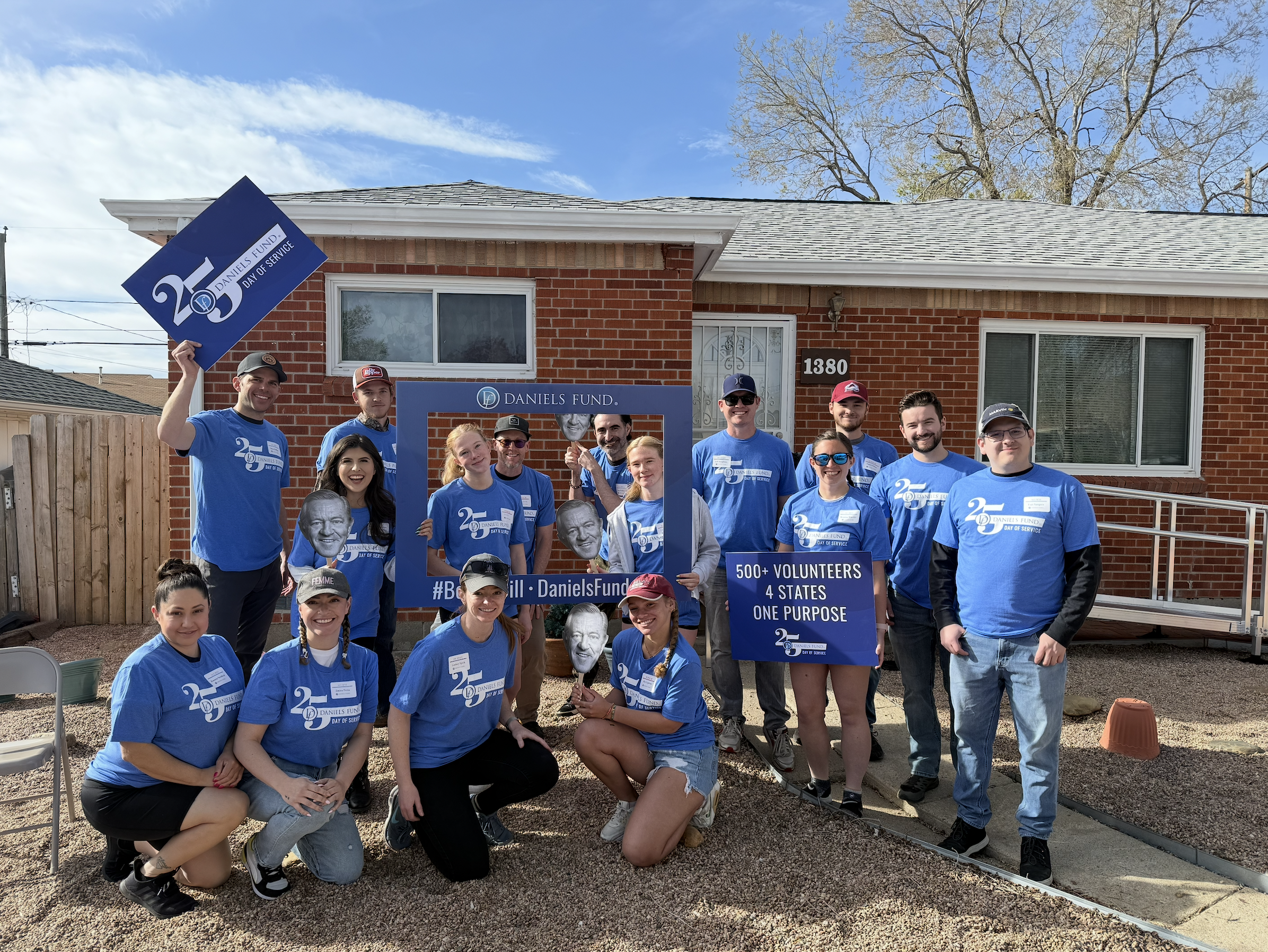 A group of volunteers in blue shirts celebrating Daniel's Fund Day of Service outside a brick house, holding signs and masks, with clear sky and a tree in the background.