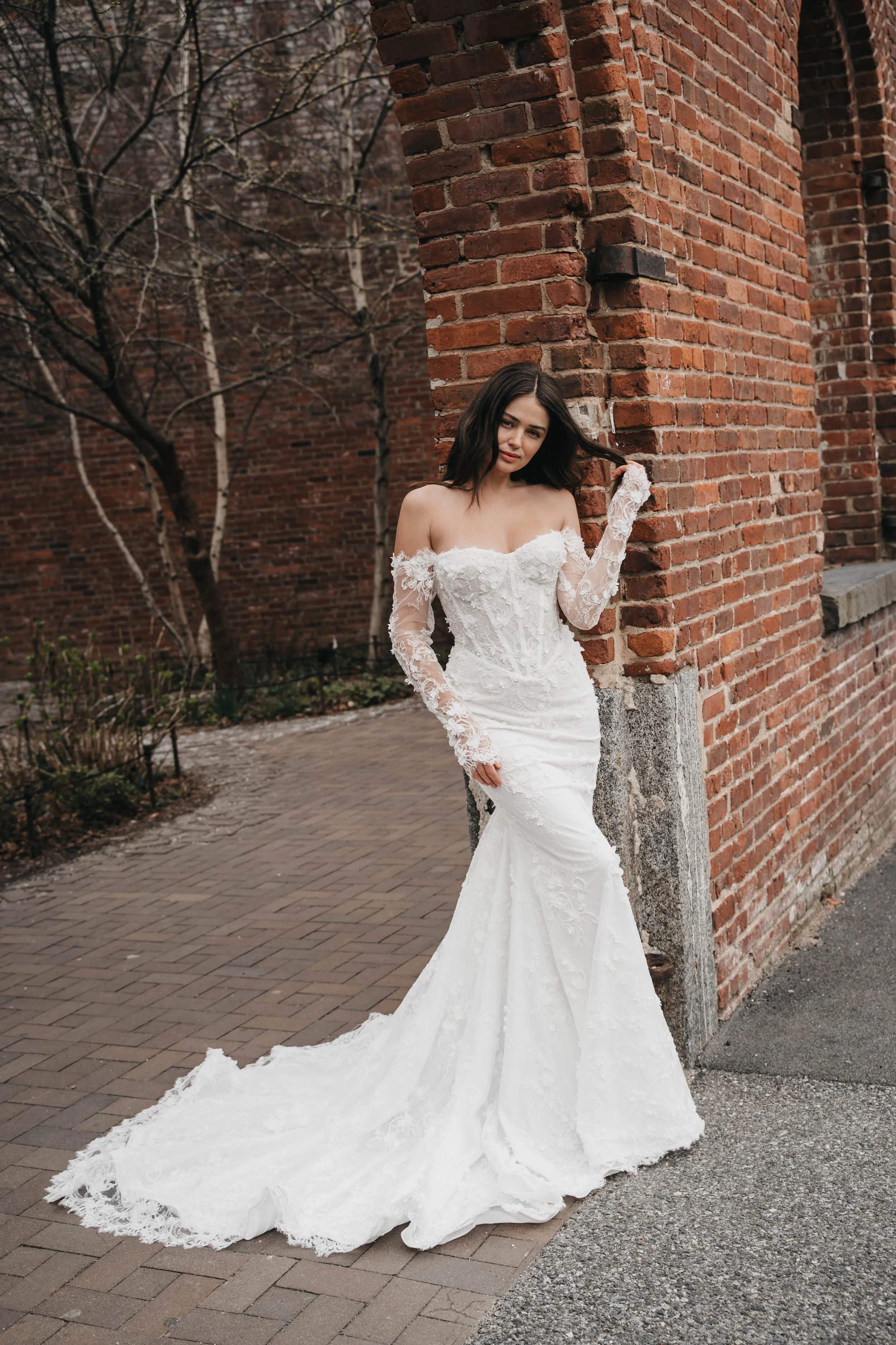 A young woman in a white lace wedding dress leaning against a brick wall outdoors, with a train extending to the pavement.