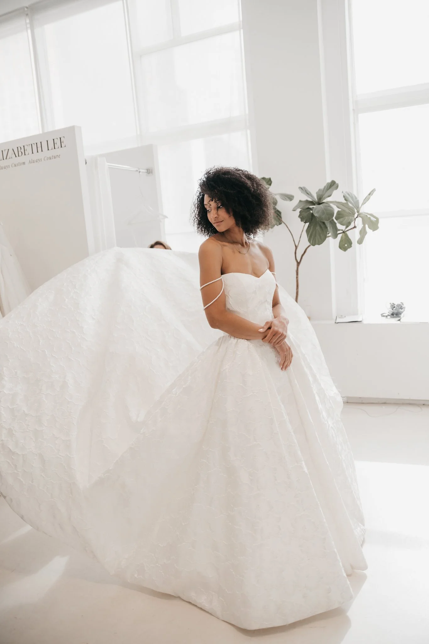 Woman wearing a wedding dress inside a bright room with large windows and a potted plant.