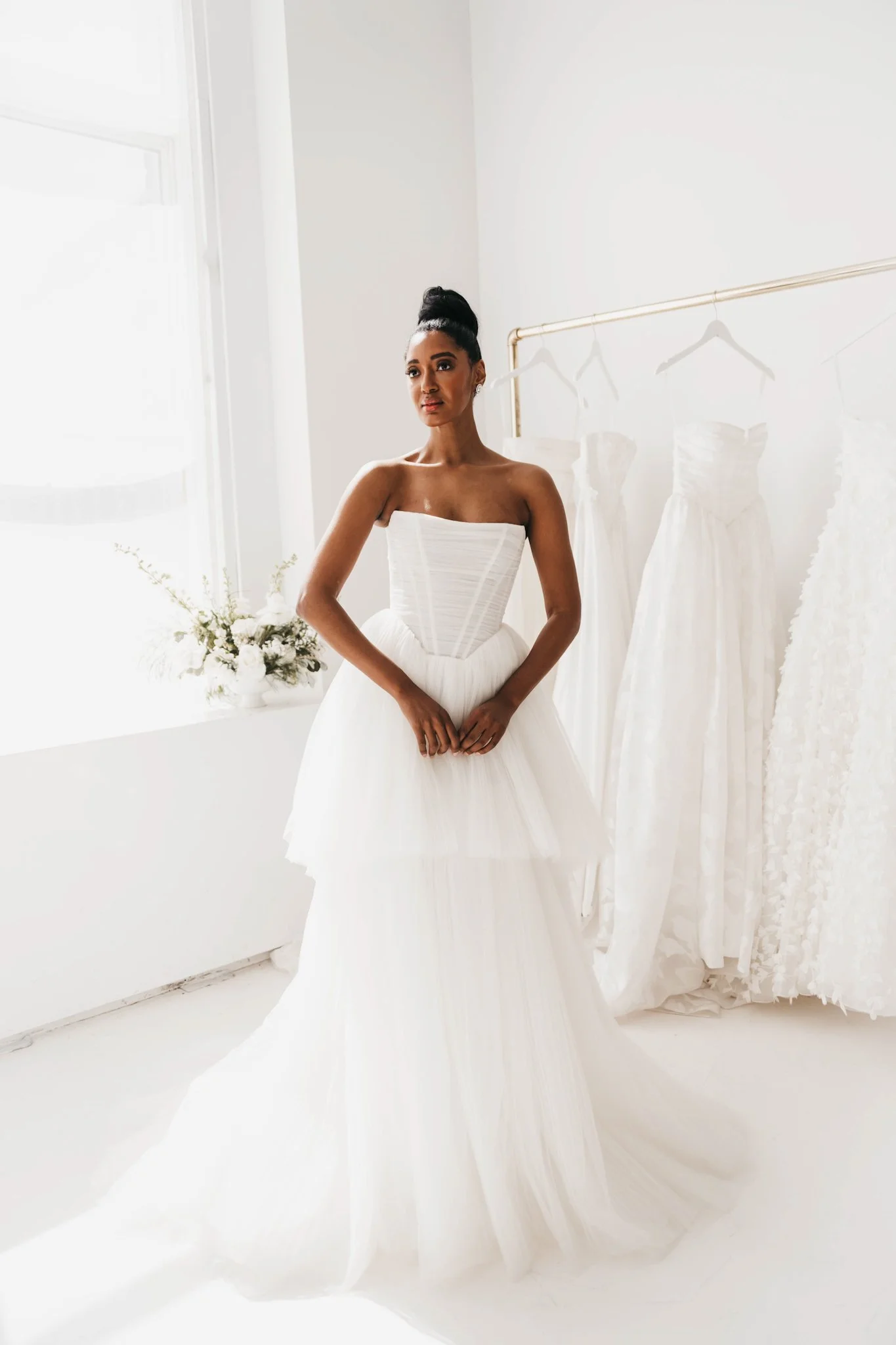 A woman in a white wedding dress standing near a window with sunlight, surrounded by several other white wedding dresses hanging on a rack and a bouquet of white flowers on a nearby table.