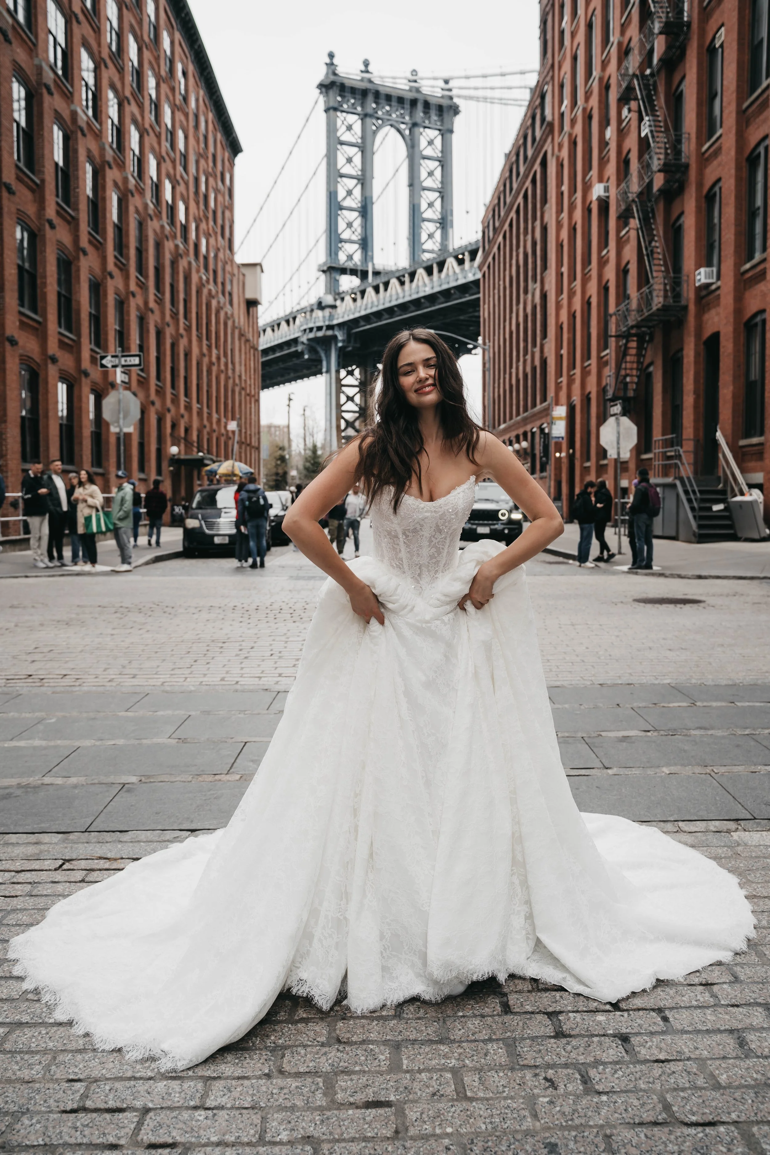 A woman in a white wedding dress standing on a cobblestone street in front of the Brooklyn Bridge in New York City.