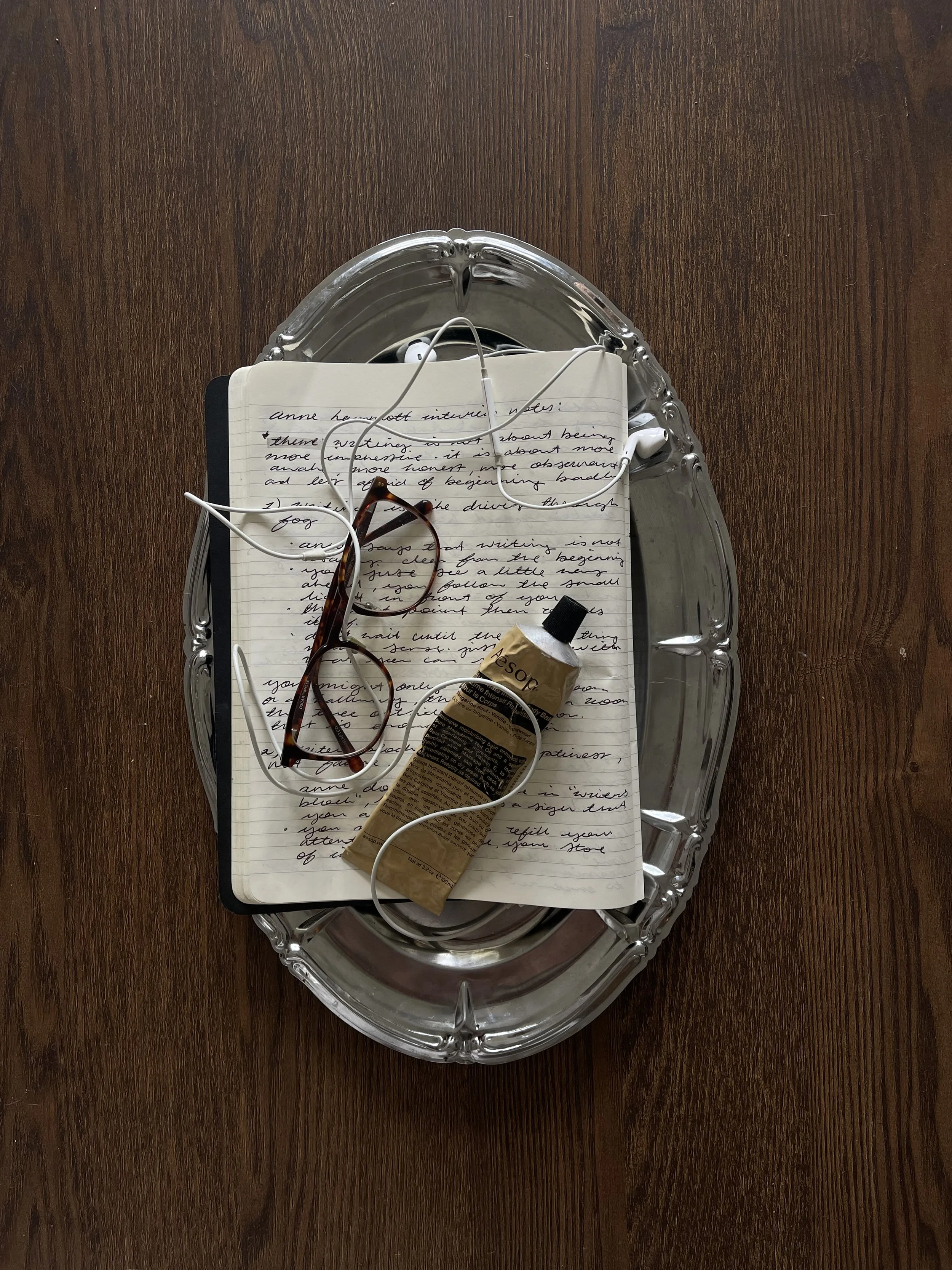 Top-down view of a silver tray with a handwritten notebook, a pair of glasses, a tube of beige cream, and white earphones on a wooden surface.