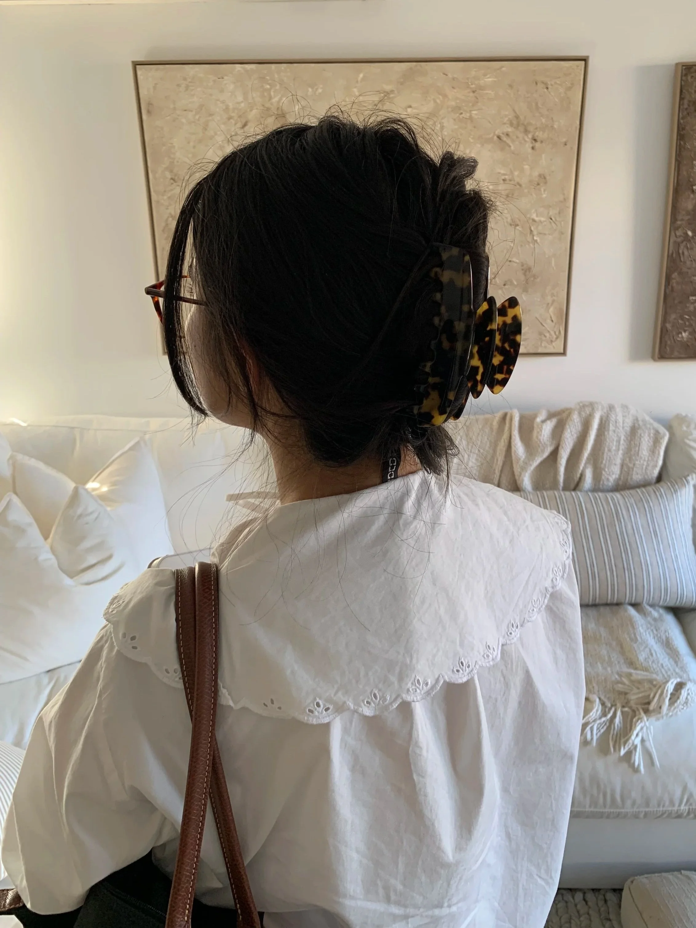 The image shows the back of a person with dark hair, wearing glasses with a tortoiseshell pattern, a white blouse with eyelet embroidery detail on the collar and sleeve cuffs, carrying a brown leather strap bag, and standing in a room with a bed, white pillows, and framed artwork on the wall.