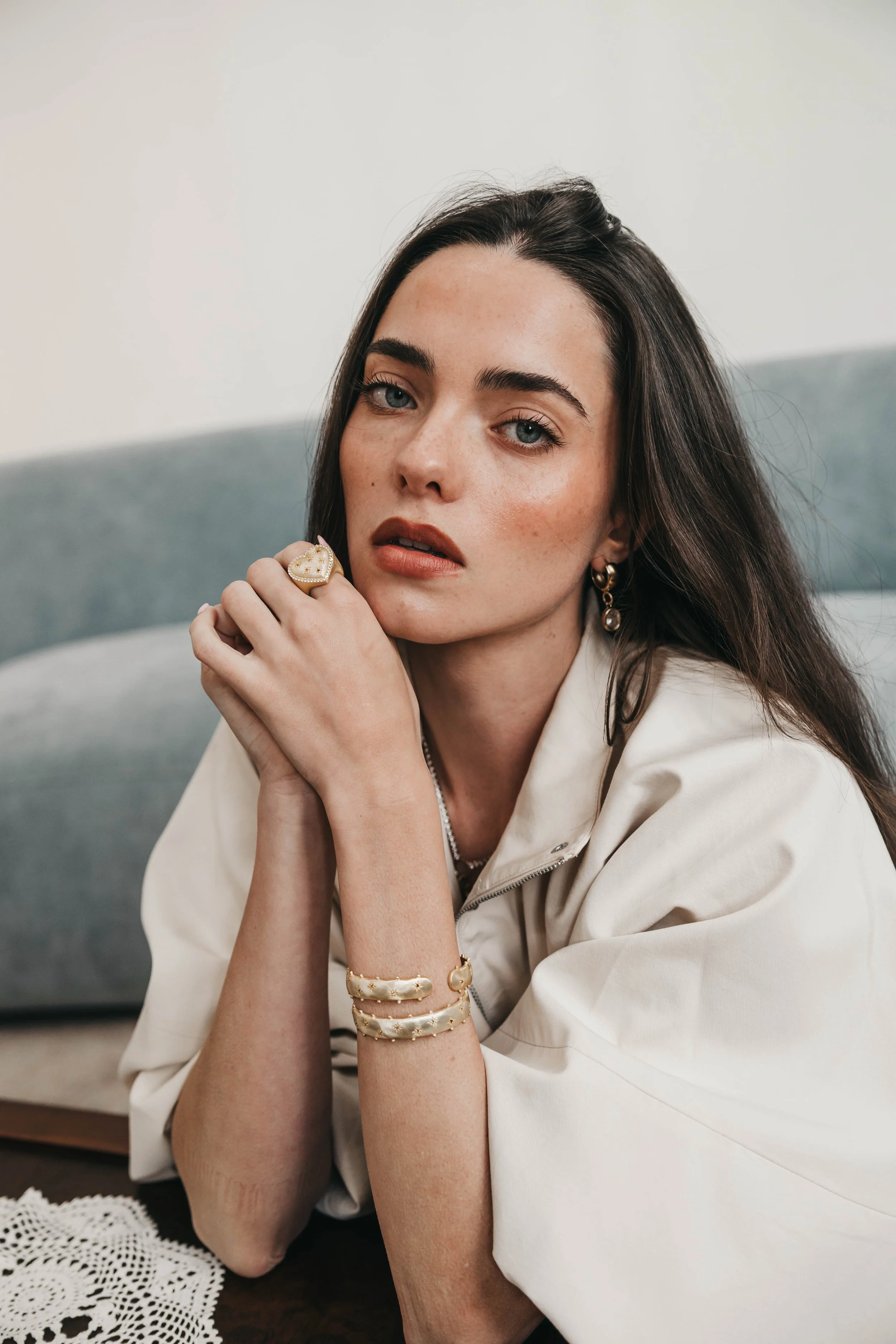 A young woman with dark brown hair and blue eyes, wearing gold jewelry including a large ring, hoop earrings, and bracelets, is resting her chin on her hand and looking at the camera with a neutral expression. She is seated at a table with a white la