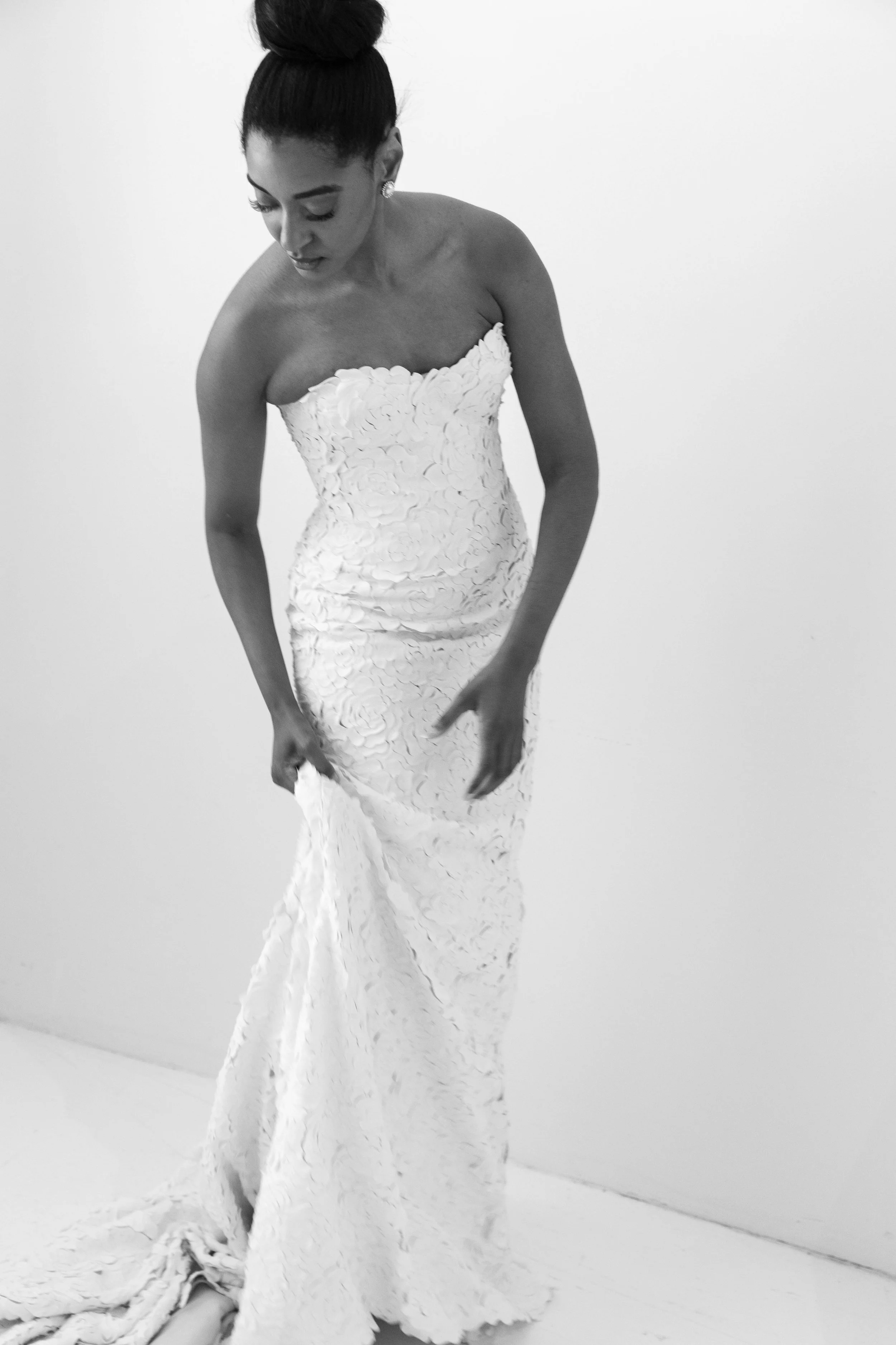 A woman in a strapless white gown with textured floral patterns, standing against a plain white background, looking downward and holding the hem of her dress.