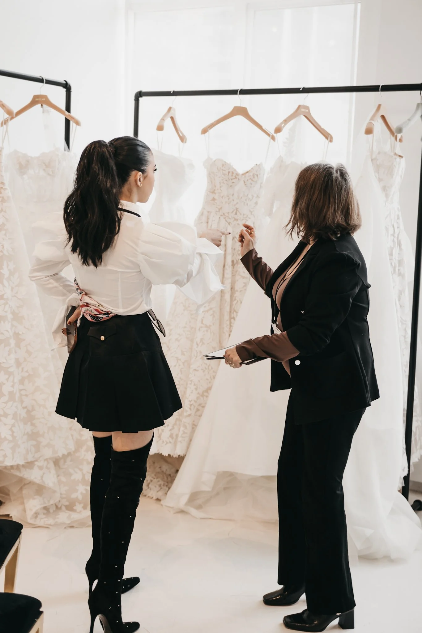 Two women are discussing wedding dresses that are hanging on a rack. The woman on the left has long dark hair in a ponytail and is wearing a black skirt and a white blouse. The woman on the right has short hair with brown highlights and is dressed in