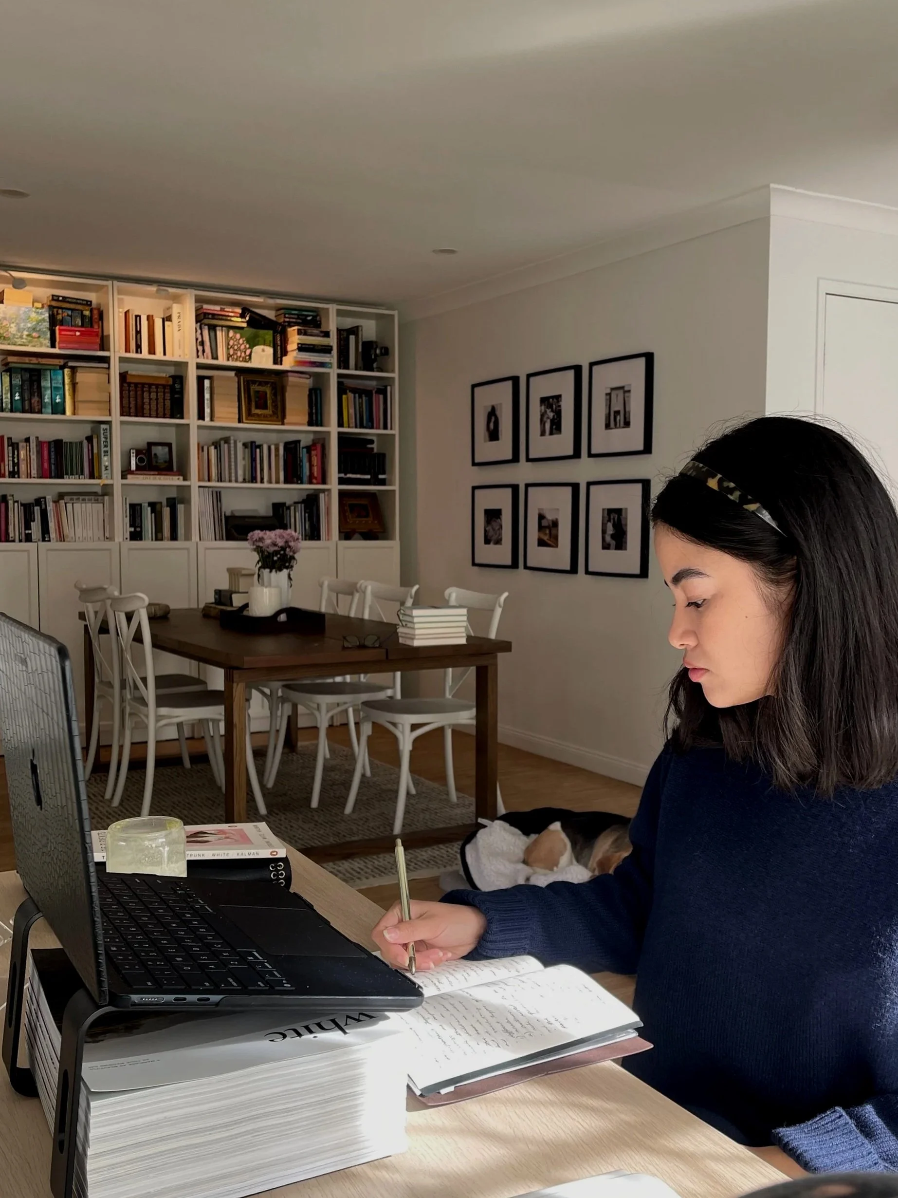 A woman with dark hair, wearing a headband, working at a desk with a laptop, surrounded by books and papers. In the background, there is a wall with framed photographs and a white bookshelf filled with books.