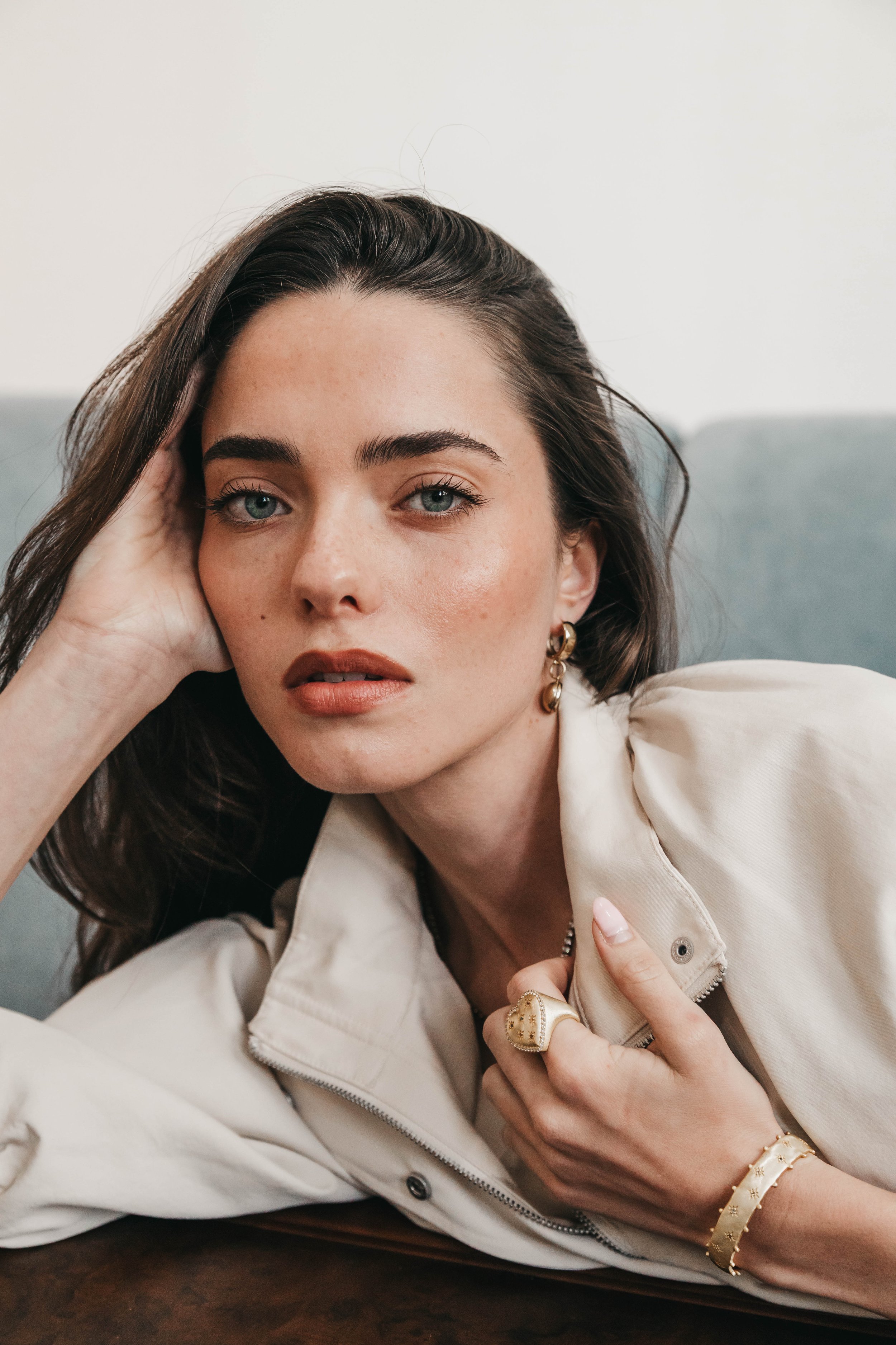 Close-up of a woman with blue eyes and dark hair, wearing gold jewelry and a beige jacket, lying on a dark wooden surface, with her hand resting on her face and her other hand touching her jacket.