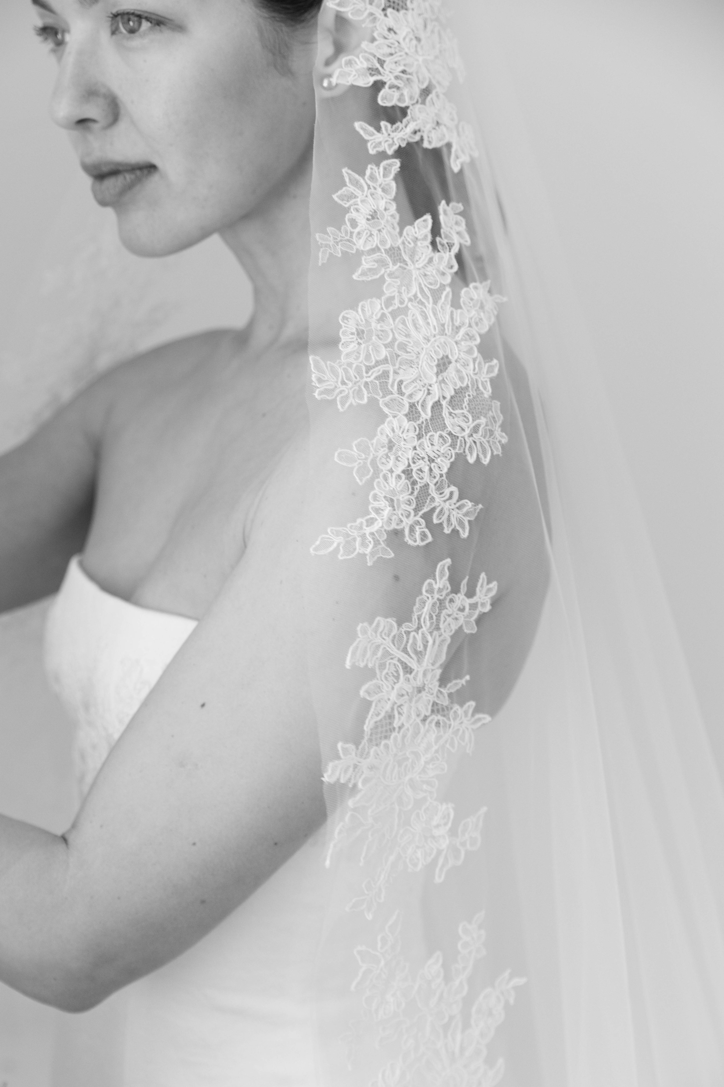 A black and white photo of a woman wearing a wedding dress and veil with floral lace embroidery, seen from the shoulders up with her face turned to the side.