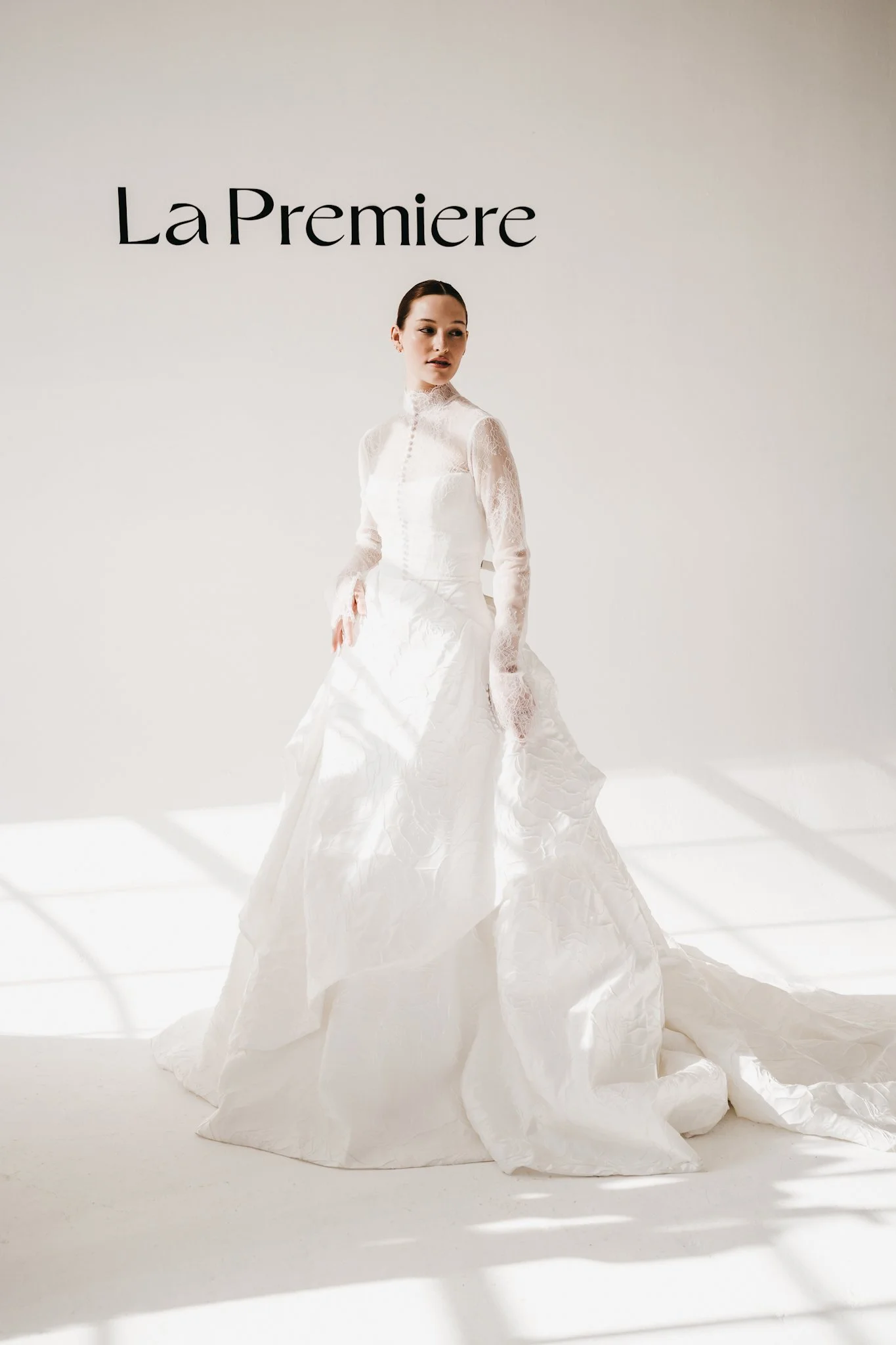 A woman in a white bridal gown standing in front of a white wall with the words "La Premiere" on it.