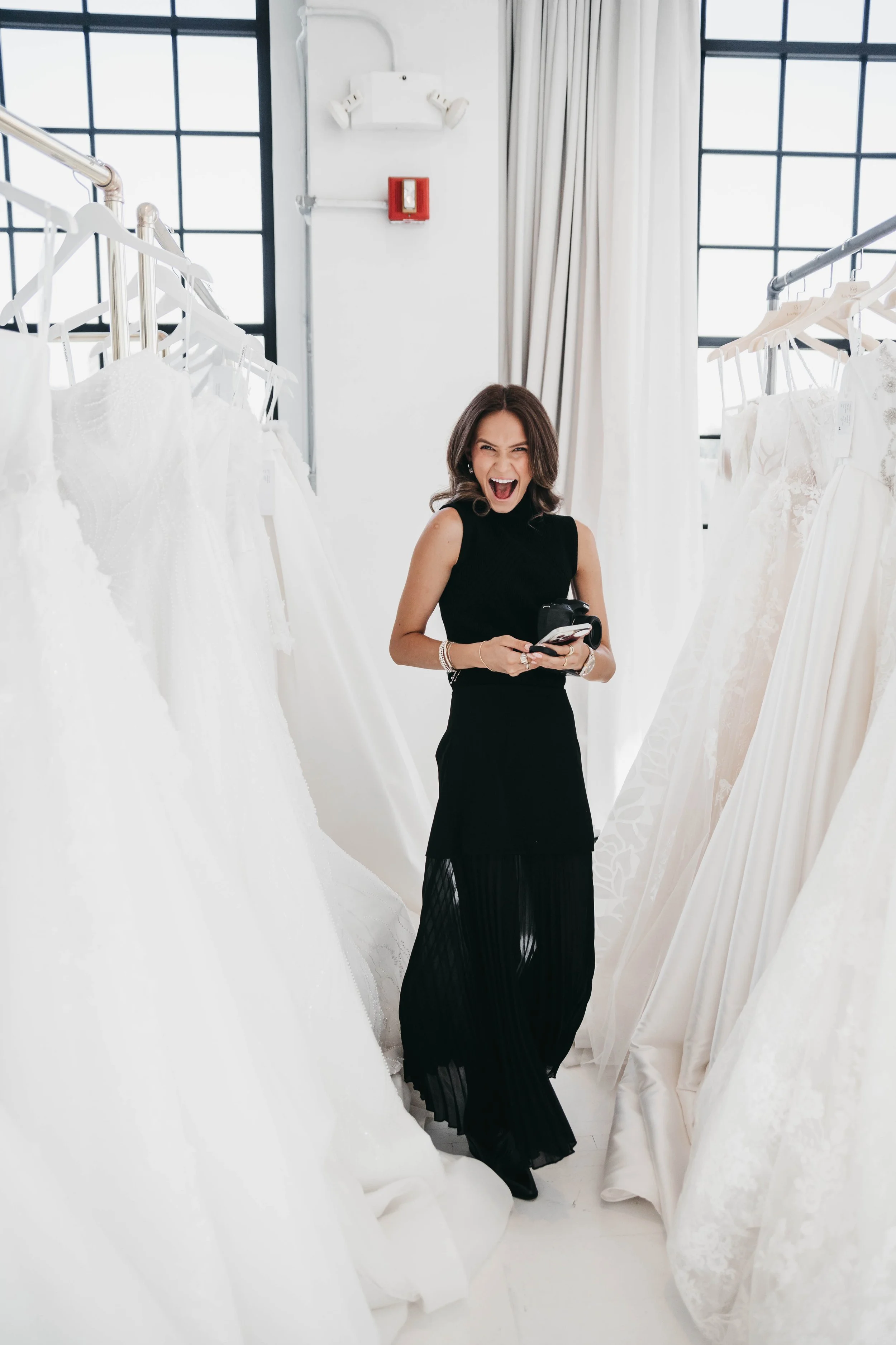 A woman with shoulder-length brown hair, wearing a sleeveless black dress and black shoes, standing in a bridal boutique surrounded by white wedding dresses on racks, smiling and holding a camera and a phone.