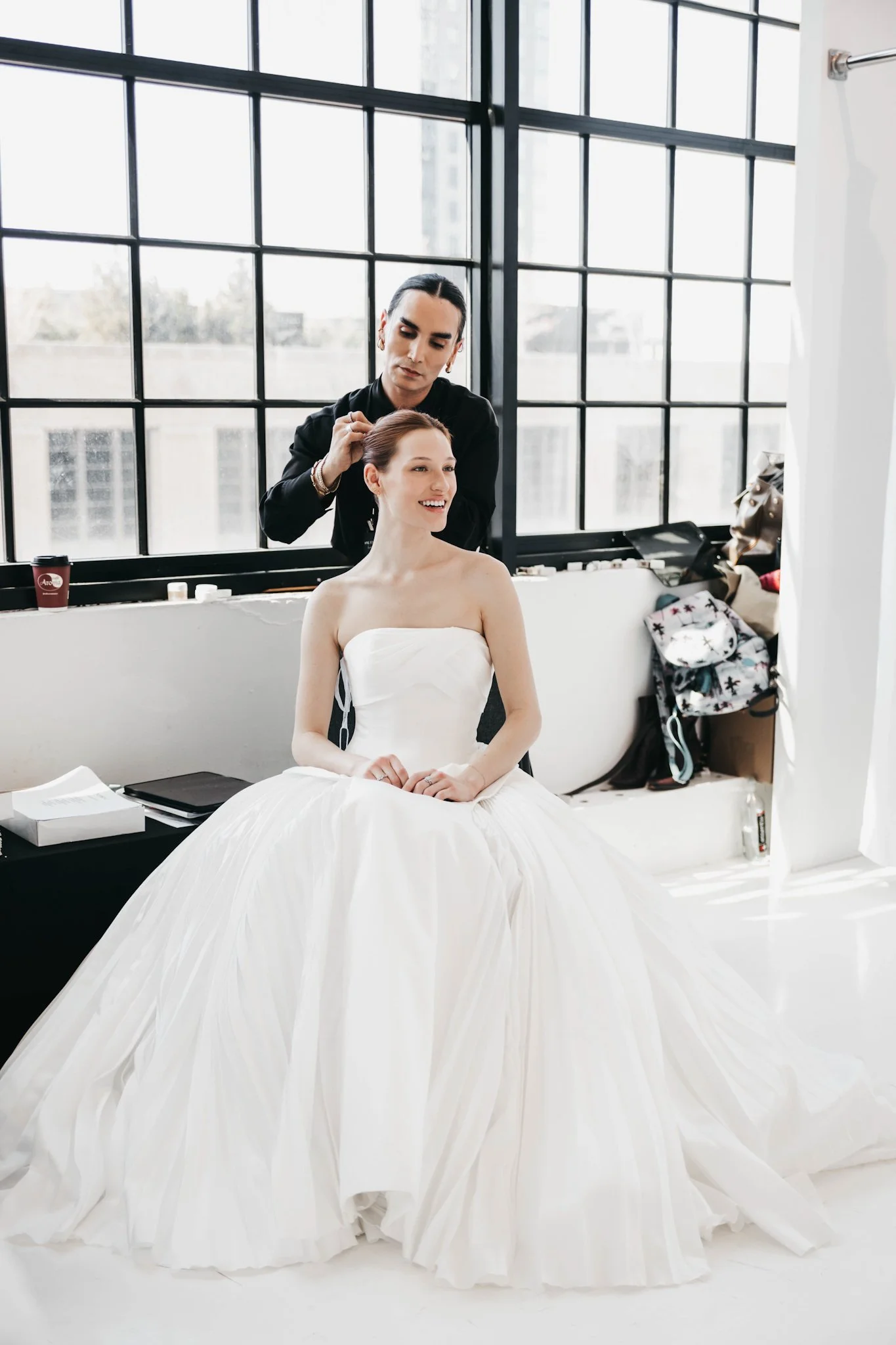 A woman in a white wedding gown smiling while sitting in a photography studio. A hairstylist standing behind her is styling her hair. The studio has large windows with city view, and various items and accessories are nearby.