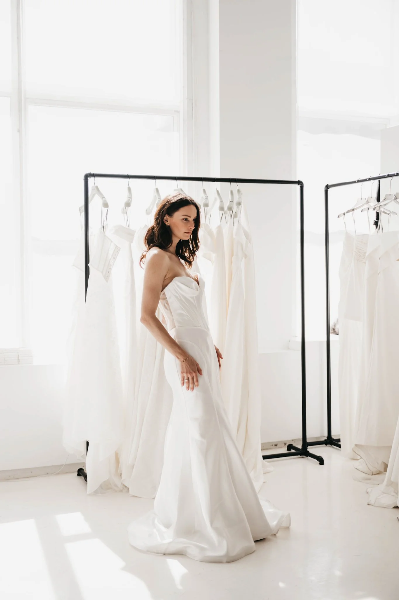 A woman trying on a white wedding dress in a bright room with large windows and hanging white dresses on racks.