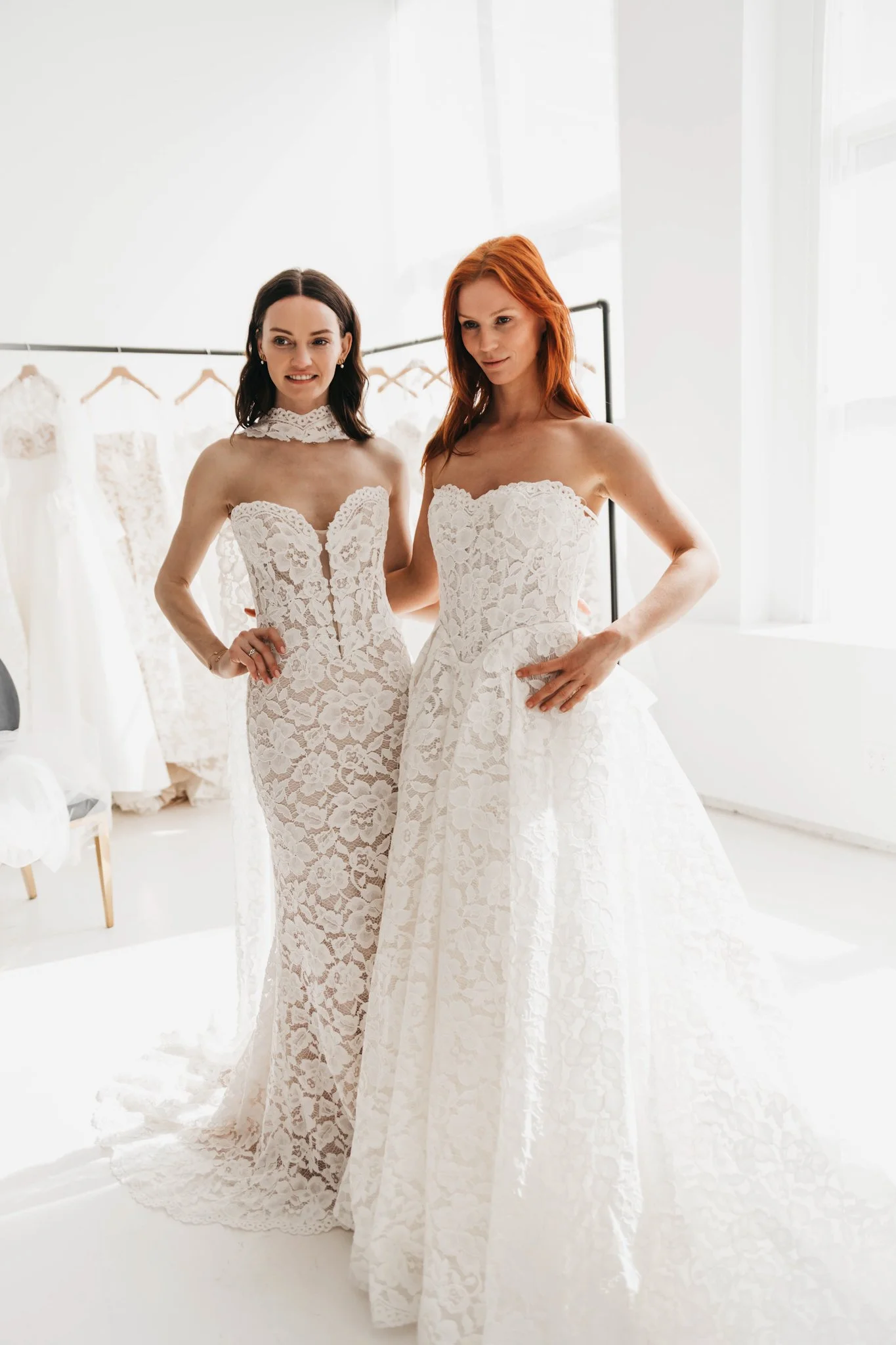 Two women trying on wedding dresses in a bright, white bridal boutique with a clothing rack of wedding gowns in the background.