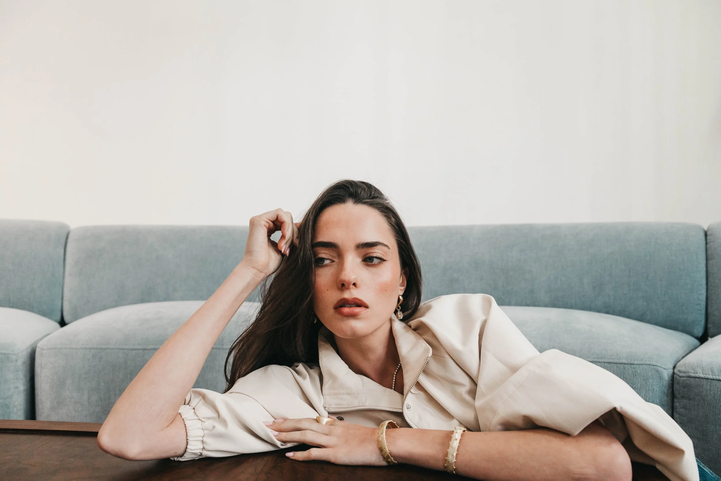 A young woman with brown hair and fair skin laying on a wooden table, propping her head with her left hand, in front of a light green sofa. She wears earrings, a beige satin shirt, and several gold bracelets, with a neutral facial expression.
