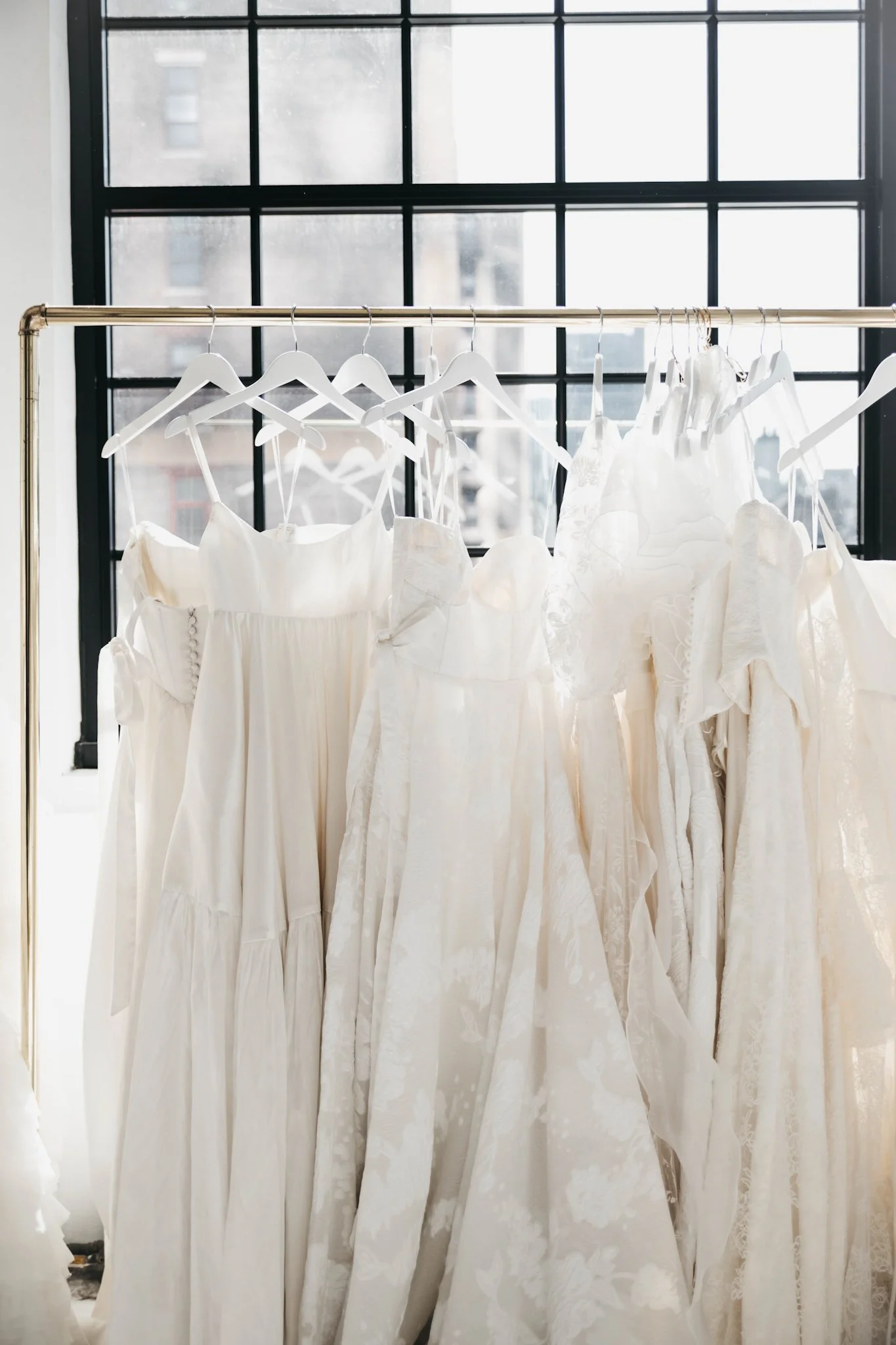 Several white wedding dresses hanging on a clothing rack in front of a large window with black window panes.