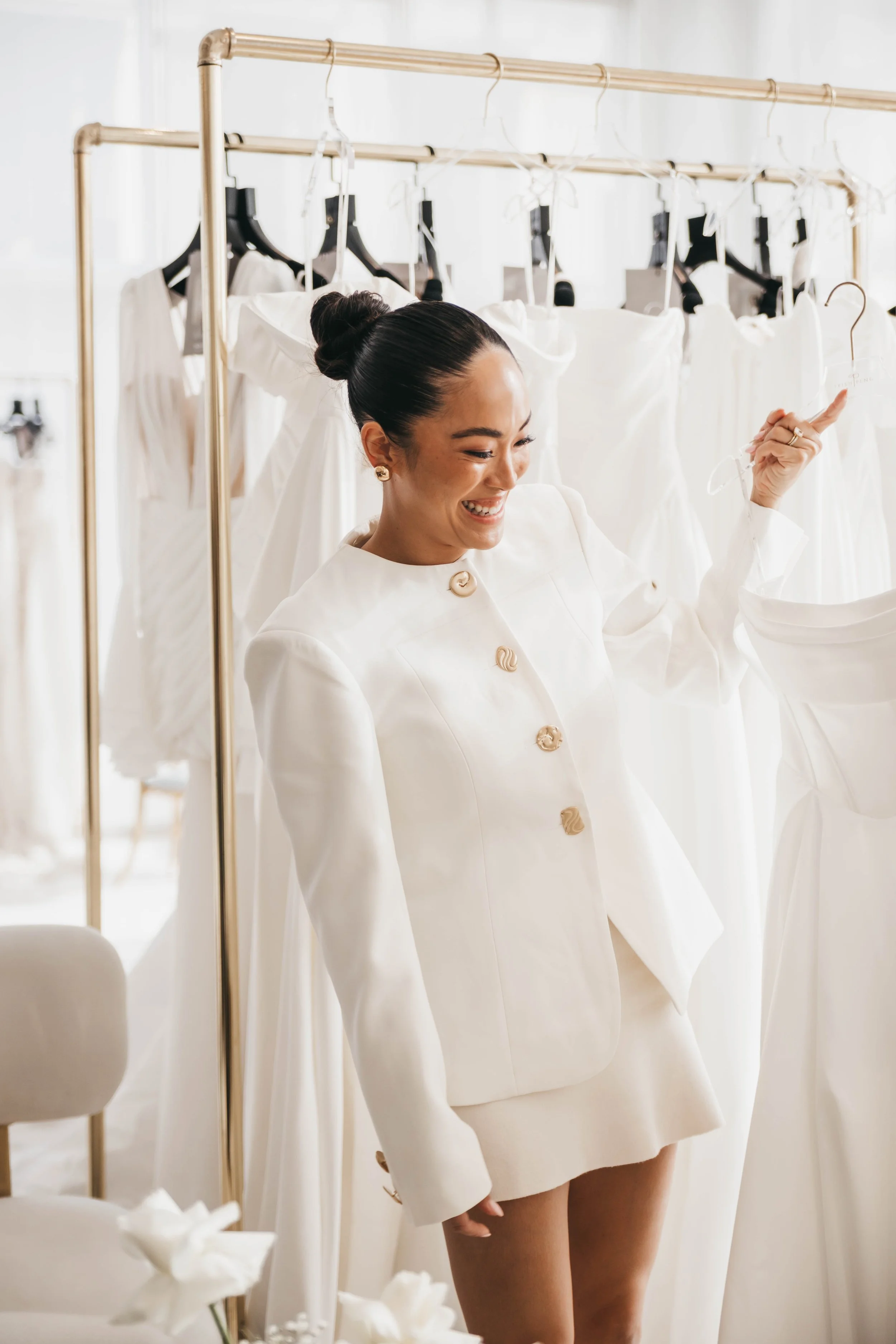 A woman smiling and pointing at white dresses hanging on a clothing rack in a bright room.