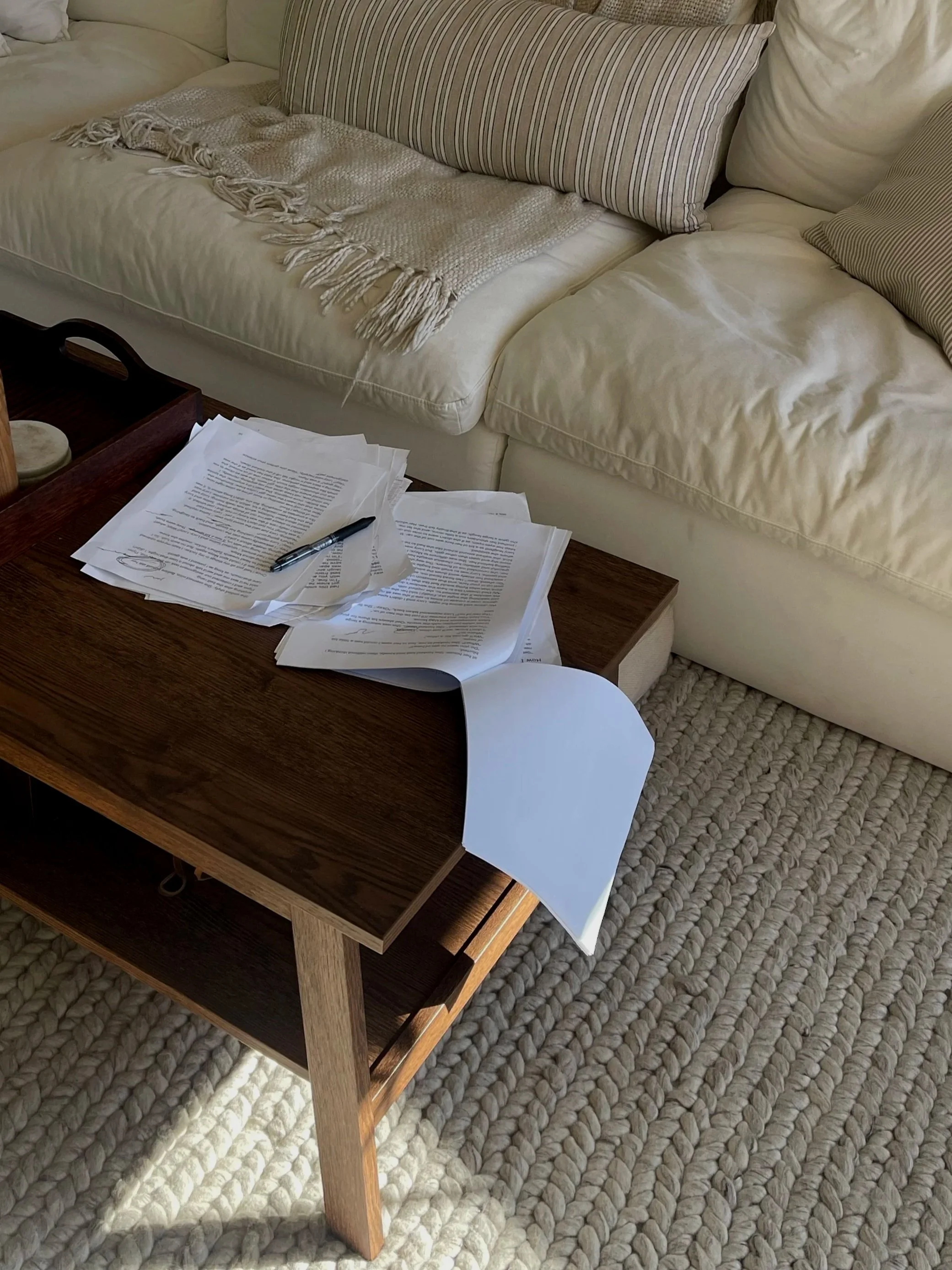 A beige sofa with striped cushions and a textured blanket, a wooden coffee table with scattered papers and a pen, and a rug on the floor.