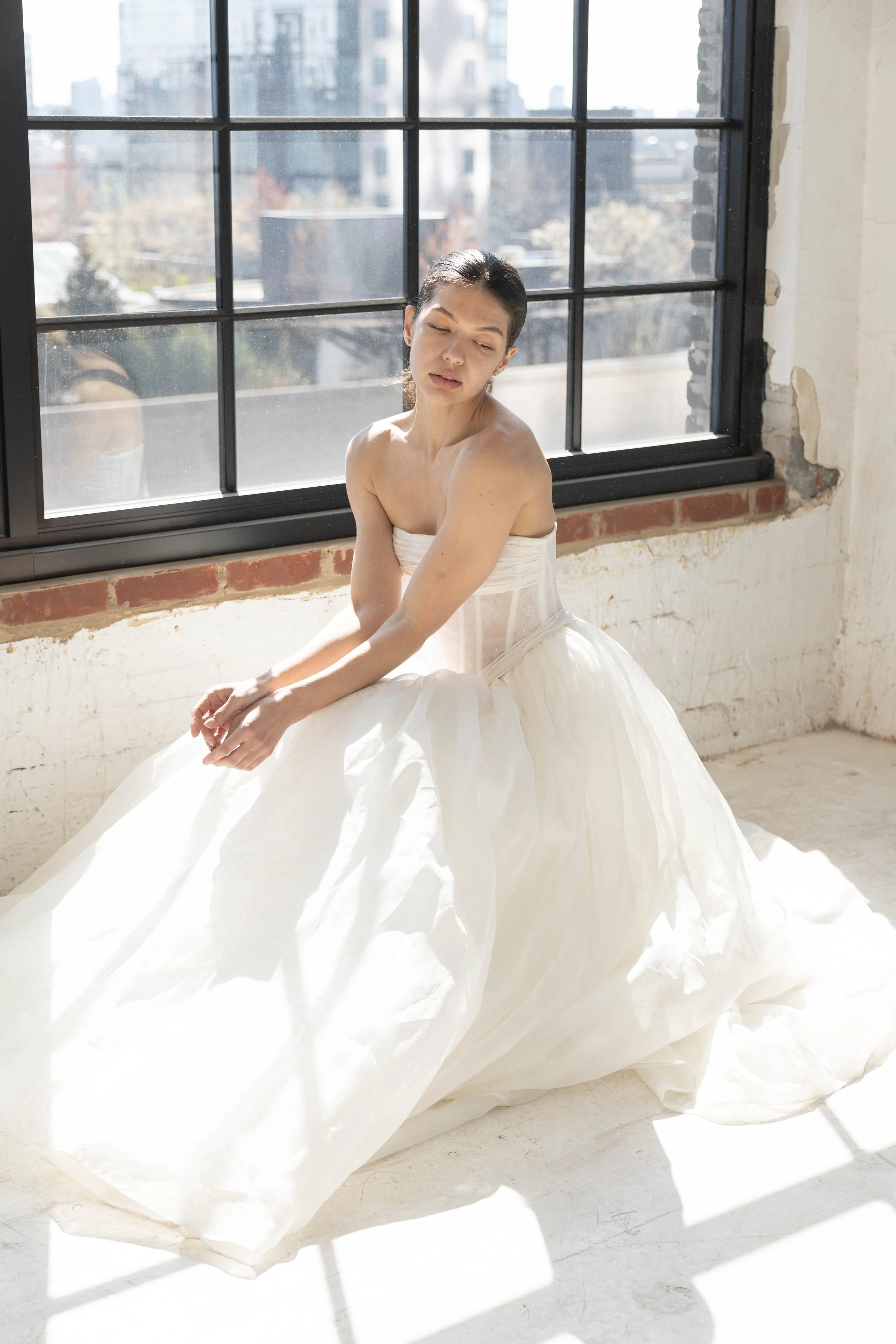 A woman in a white dress sitting near a large window with black frames, city buildings visible outside, soft sunlight illuminating her and the style of the dress appears formal and elegant.