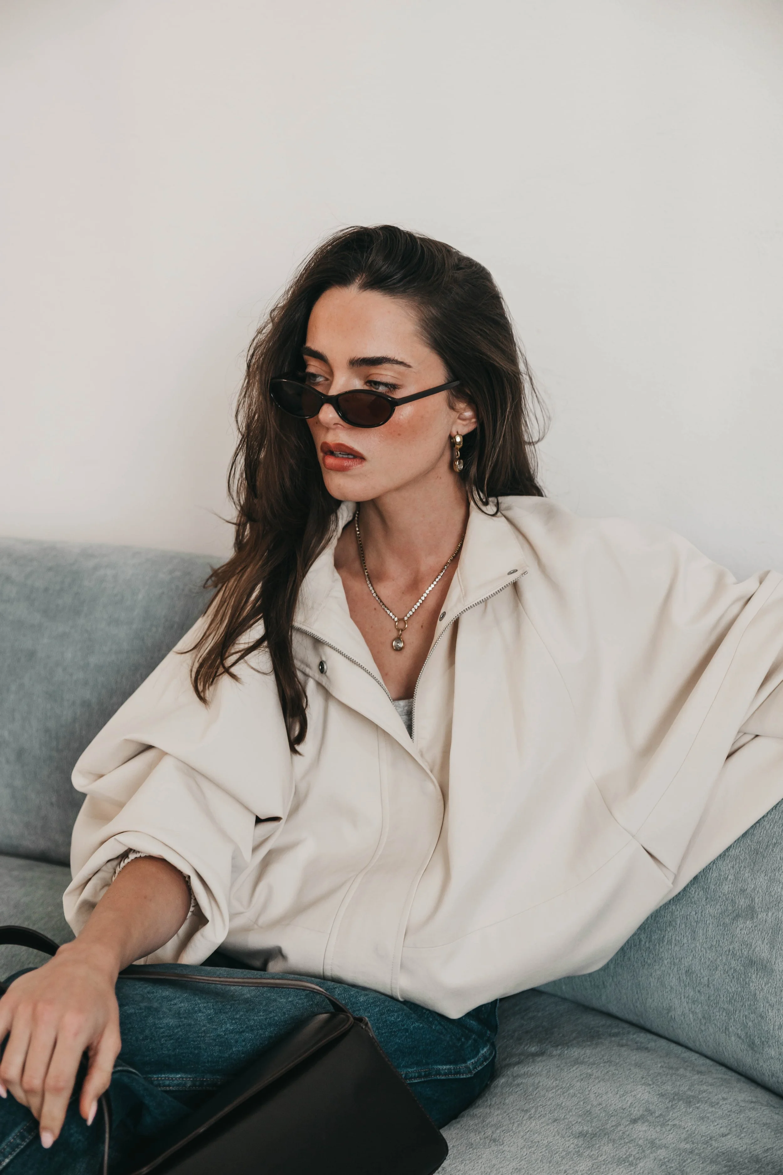 A woman with long dark hair wearing black sunglasses, gold earrings, and layered necklaces, dressed in a white jacket and blue jeans, sitting on a gray sofa against a plain white wall.