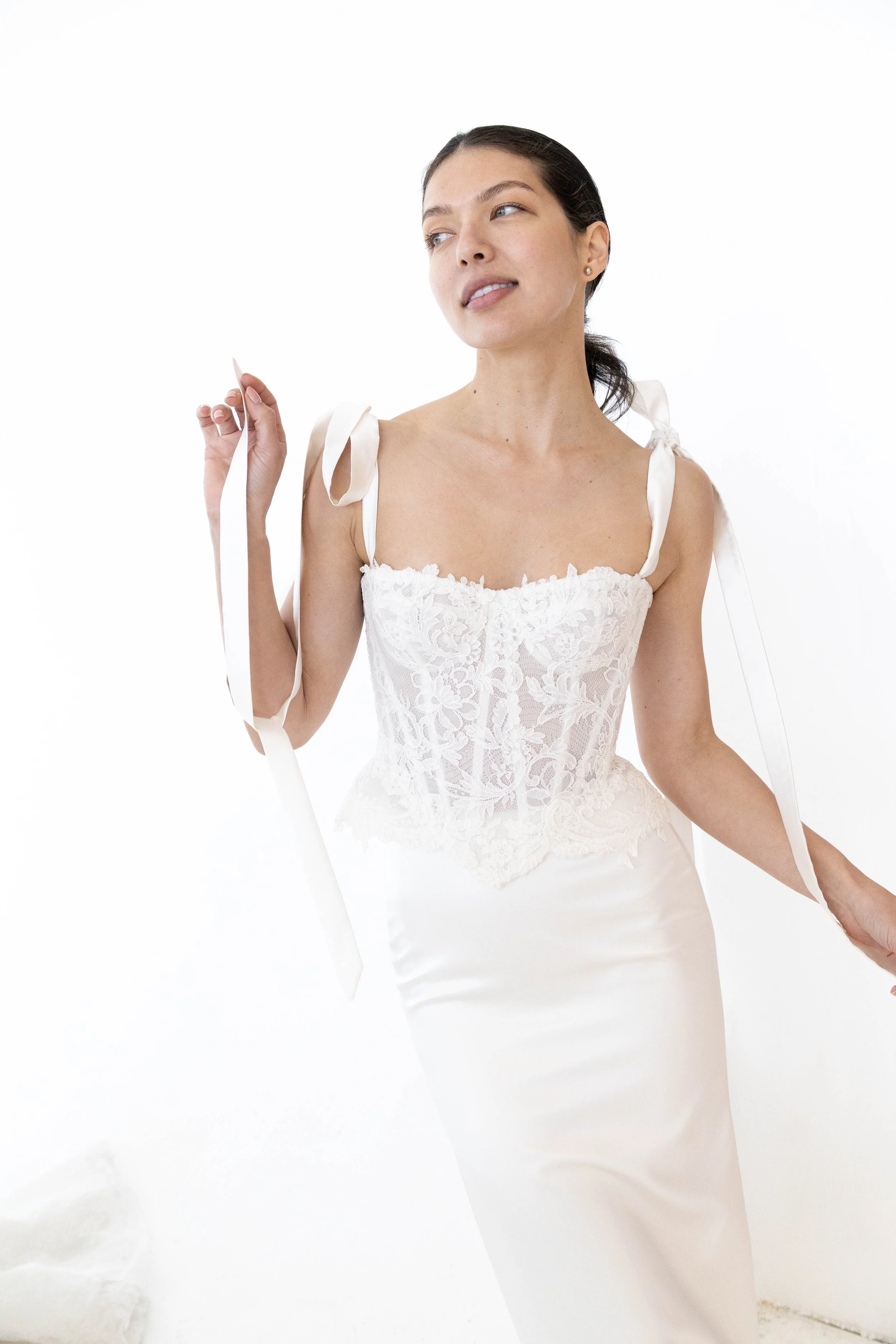 A woman wearing a white lace dress with satin straps, standing against a plain white background, holding a piece of ribbon.