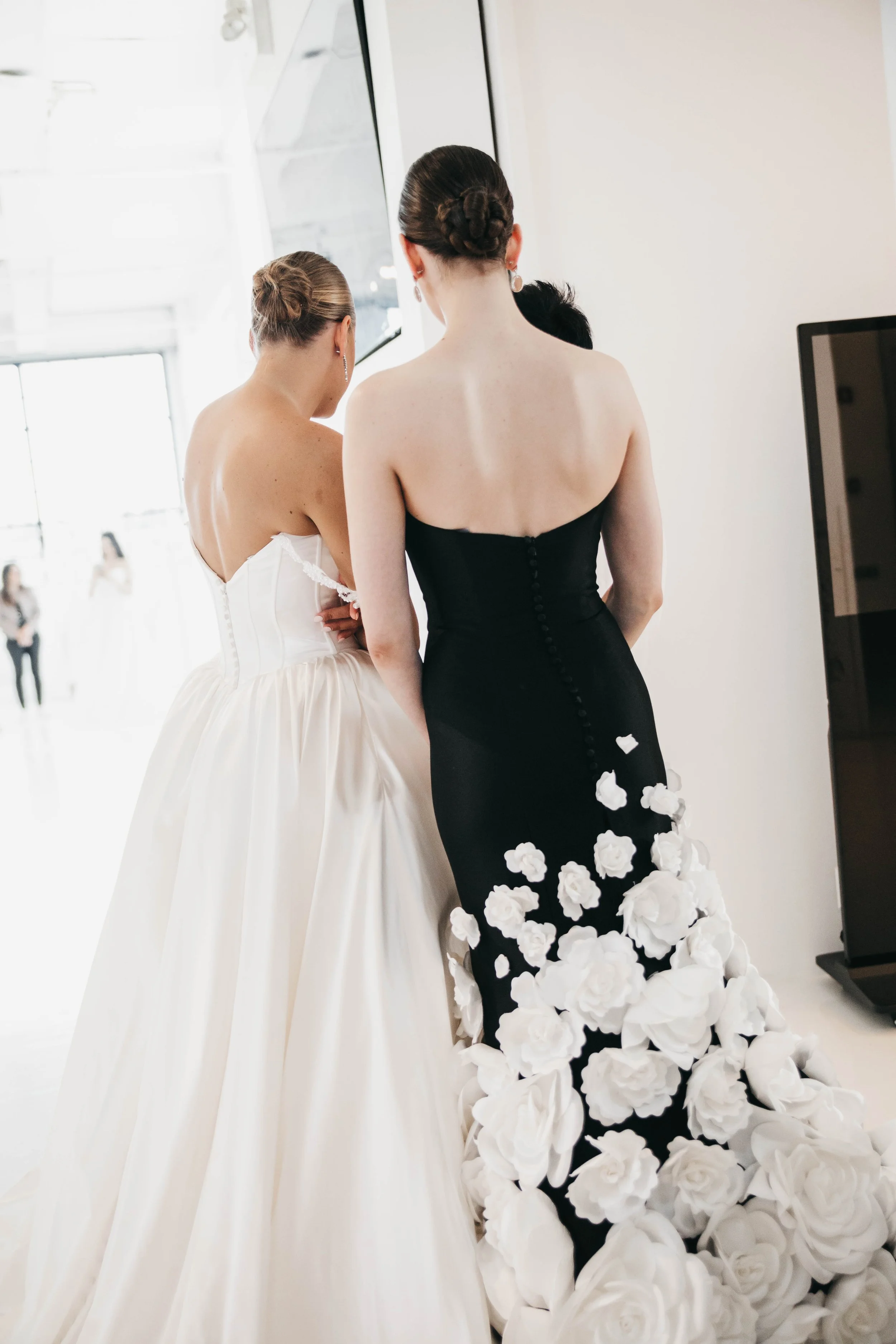 Two women in formal dresses, one in white and one in black with white floral details, standing with their backs to the camera in an indoor setting, possibly preparing for a wedding or special event.
