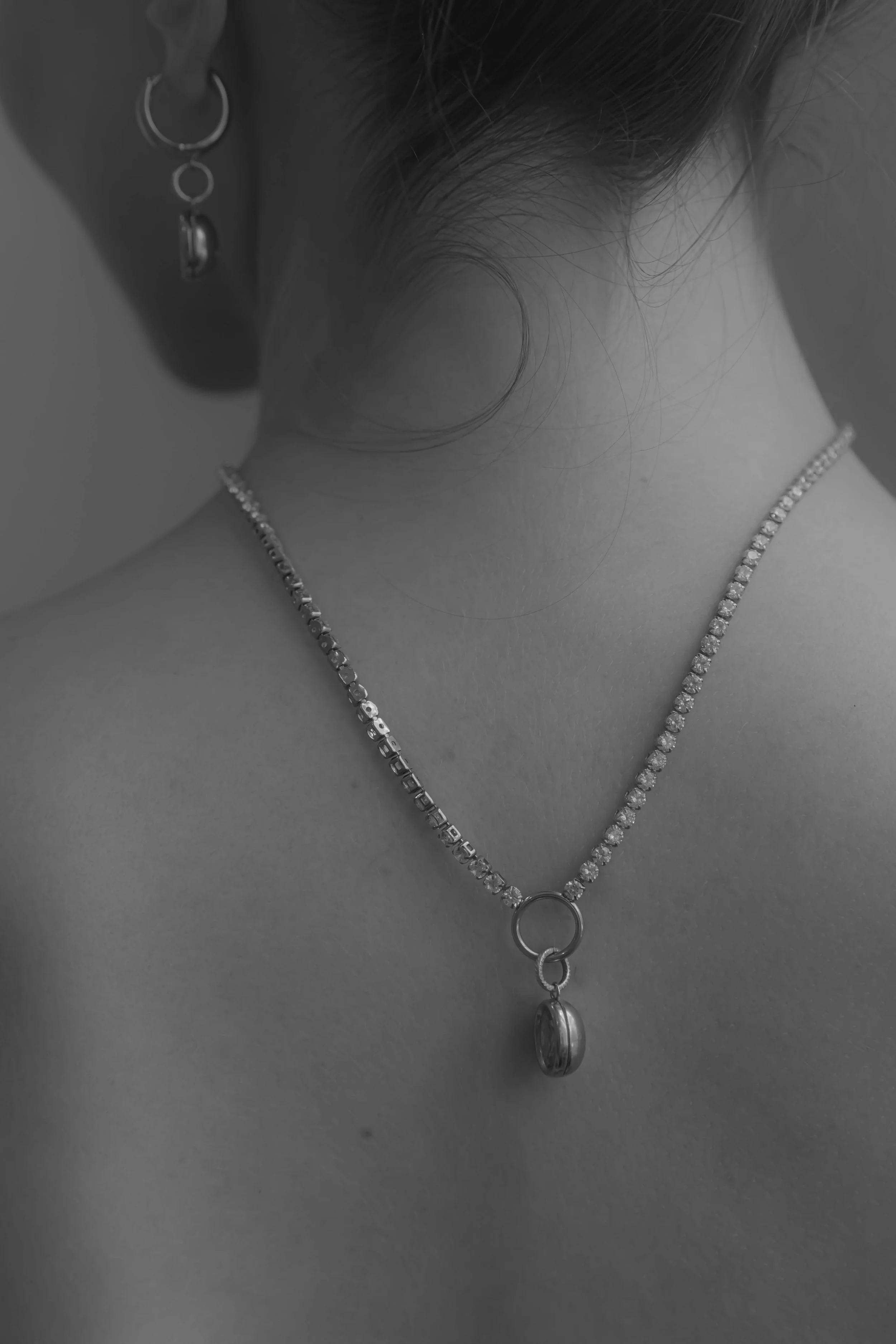 Close-up of a person's neck and upper shoulders, wearing a diamond necklace with a large pendant and earrings with circular and hoop designs, in black and white photography.