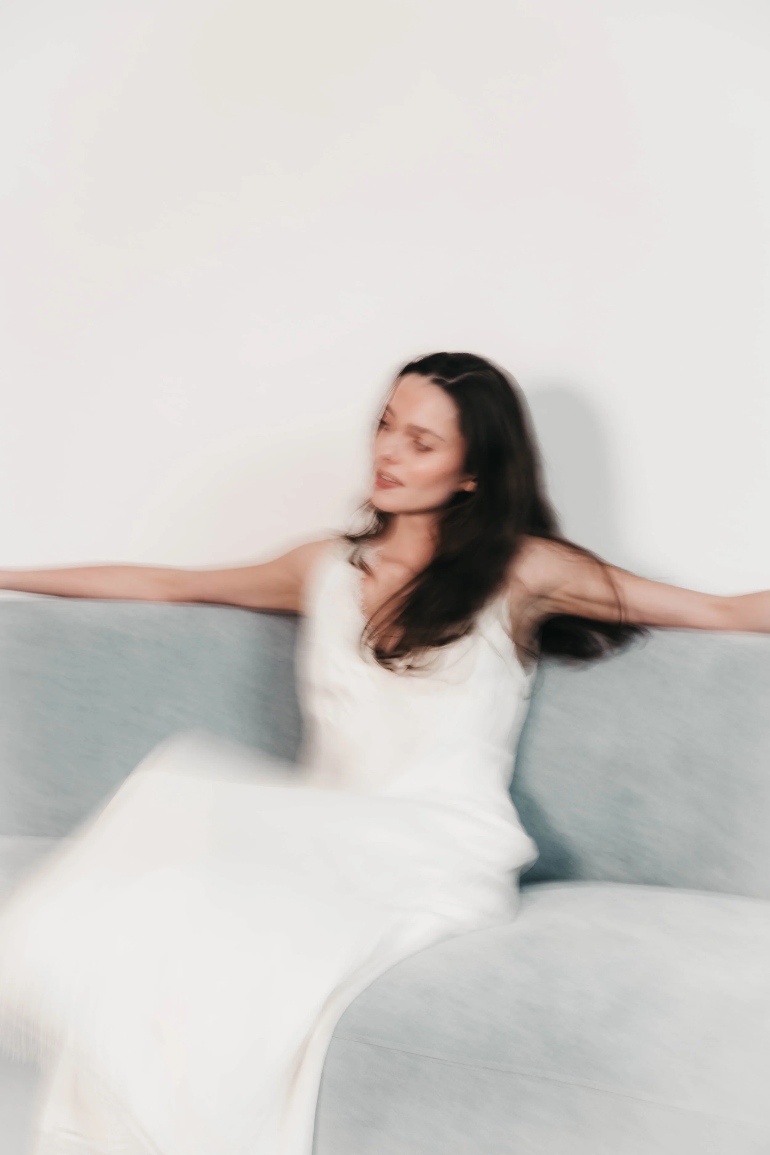 A woman with long dark hair wearing a white dress, sitting on a sofa with arms outstretched, against a plain white wall.