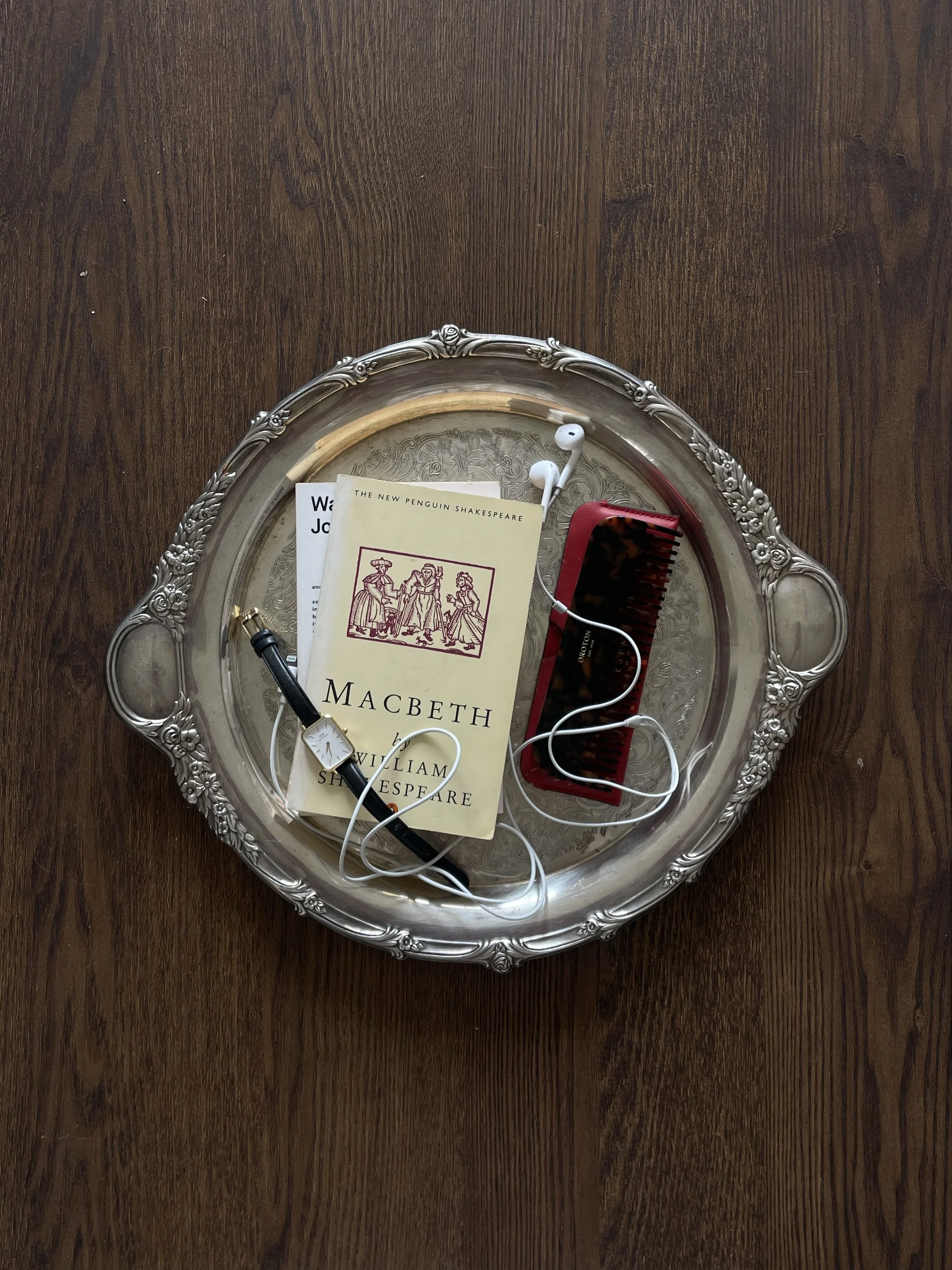 A silver tray on a wooden surface holding a yellow book titled 'Macbeth' by William Shakespeare, with white earphones, a red hairbrush, and a black pen, along with some papers.