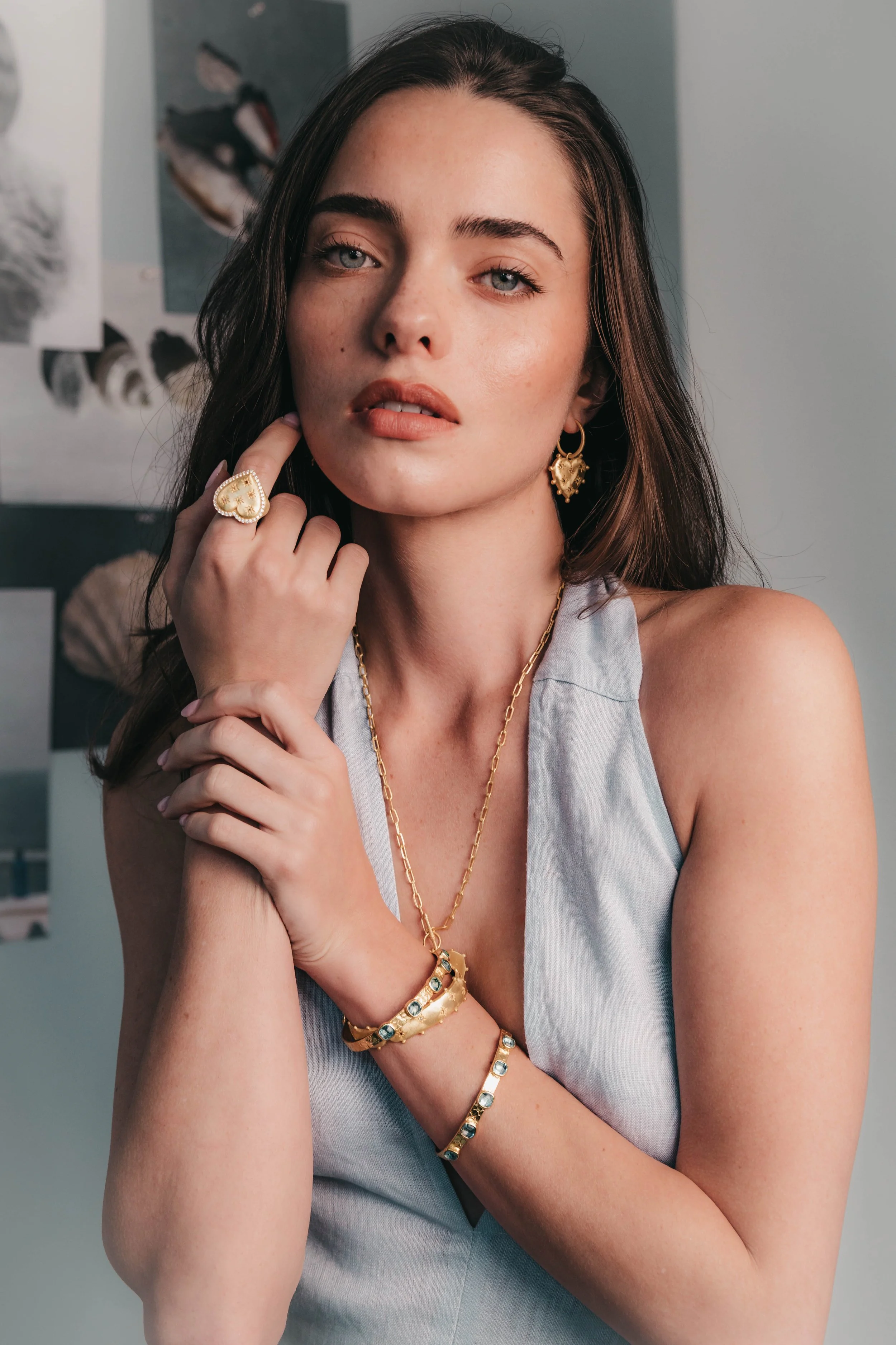 Close-up of a woman with dark hair and blue eyes, wearing vintage jewelry including a large heart-shaped ring, heart-shaped earrings, and multiple gold bracelets and necklaces. She is holding her chin lightly with her left hand and looking directly a