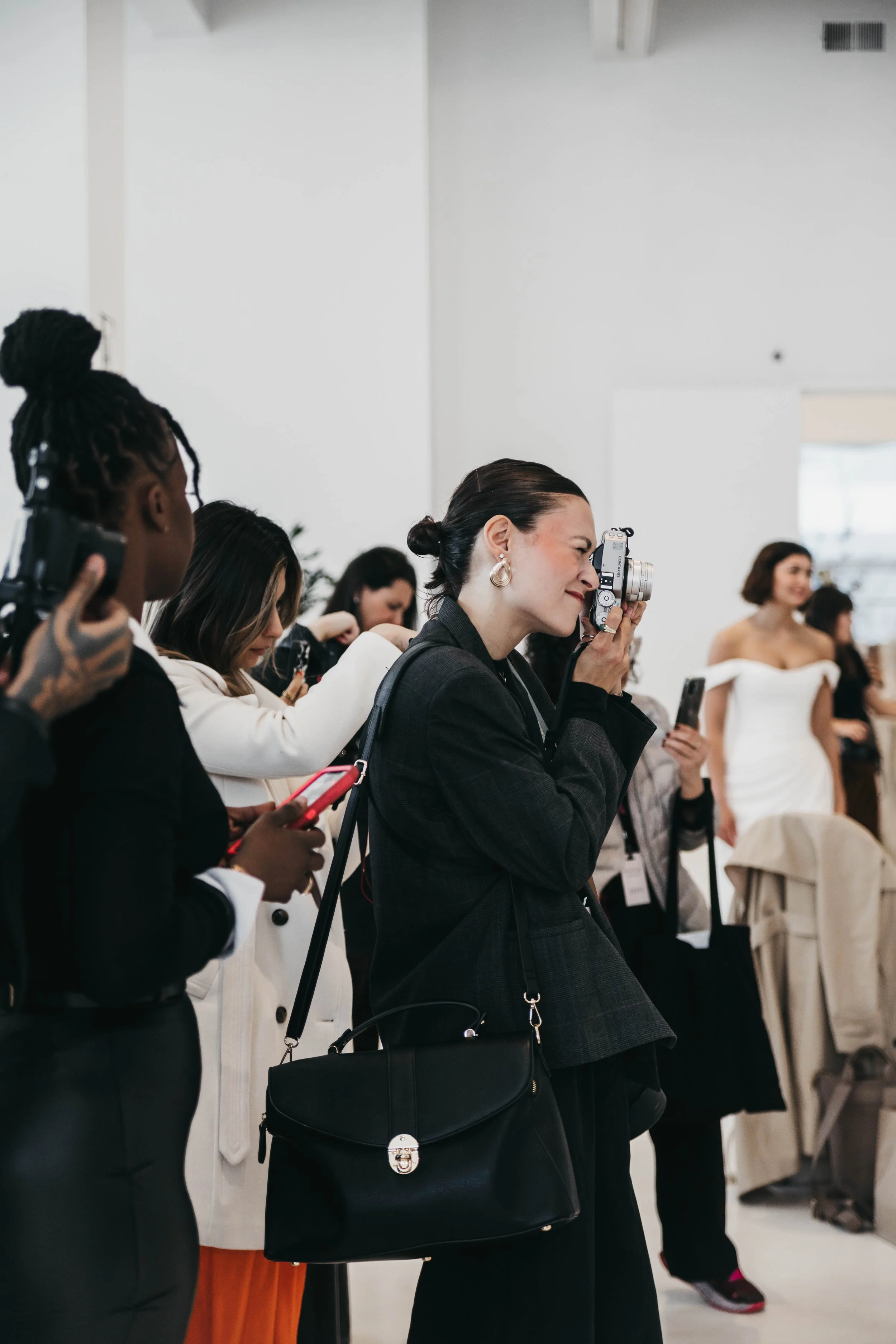 Women attending a formal event, with one woman taking photos with a camera, others using mobile phones, in a bright room with white walls and chairs.